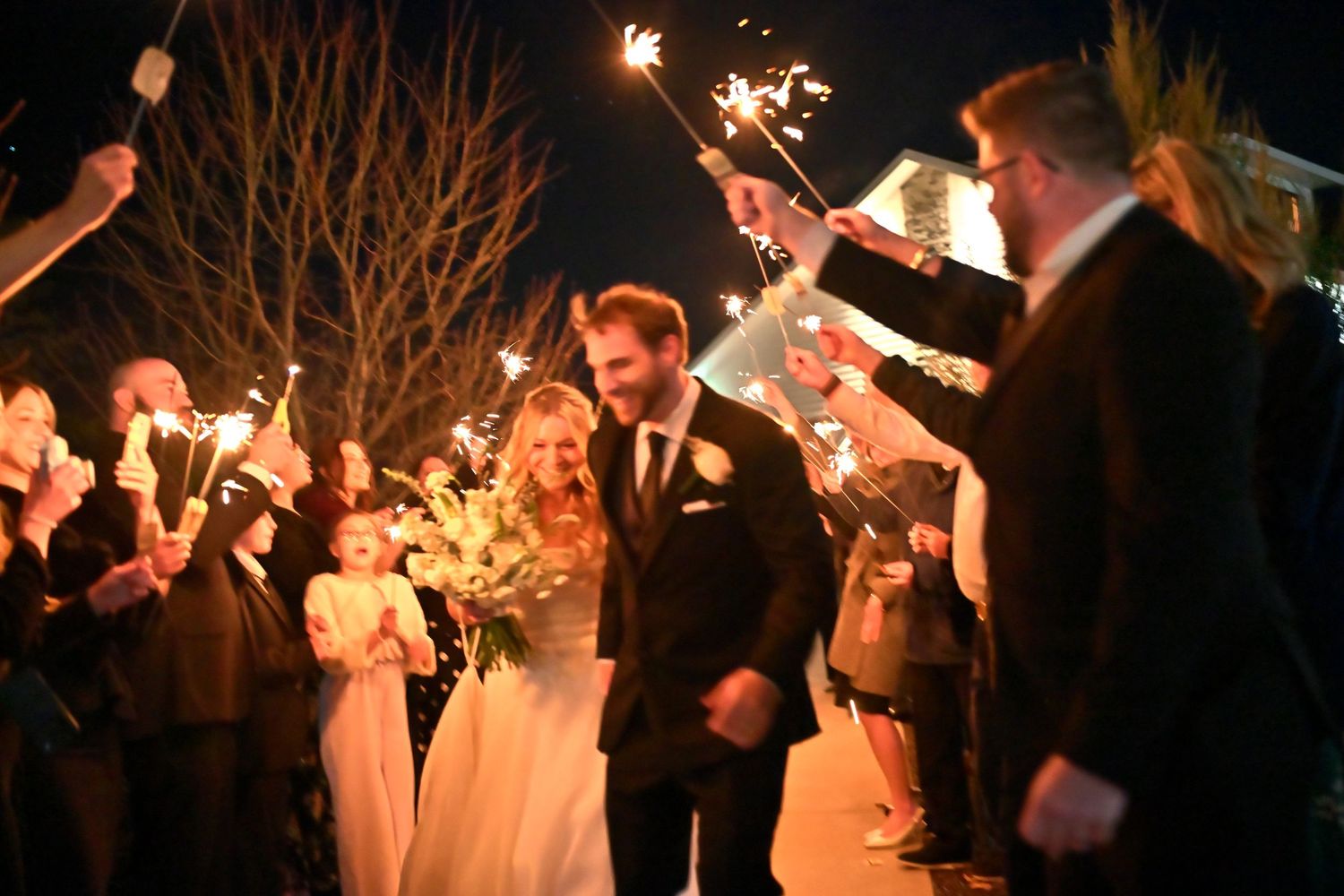 Bride and groom walk through sparkler exit surrounded by wedding guests at night.
