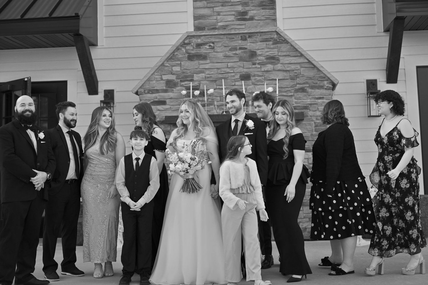 Black and white wedding party photo outside a chapel with bride, groom, and guests in formal attire.