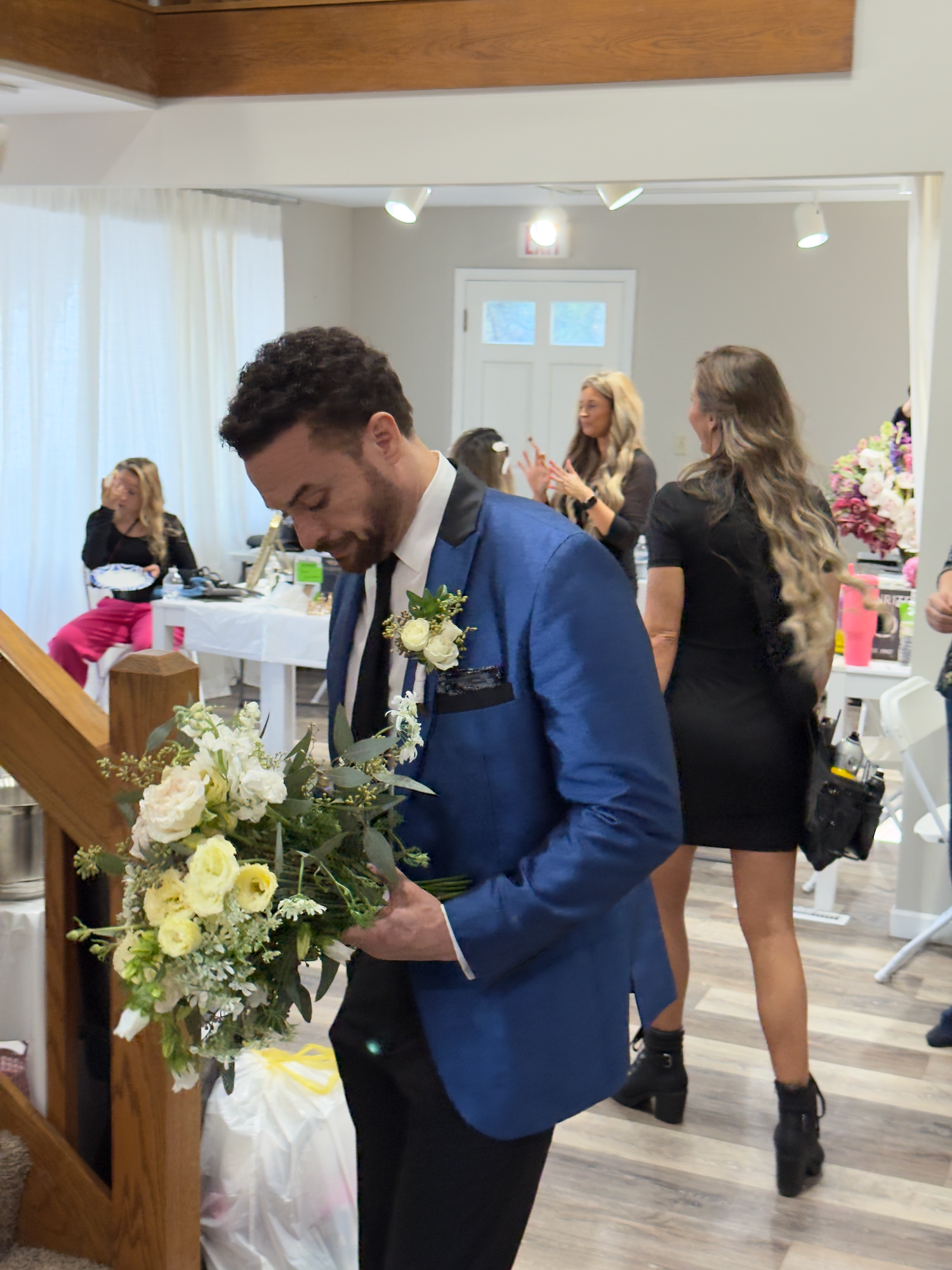 Wedding guests gather in a bright reception space with white decor and floral arrangements in blue and cream colors.