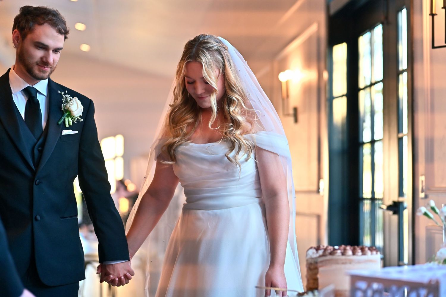 Bride in white gown and veil holds hands with groom in dark suit at elegant wedding reception venue.