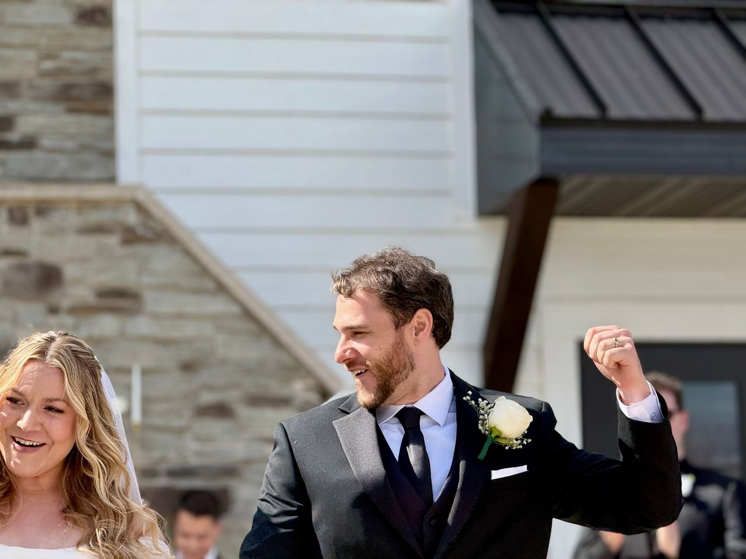 Joyful newlywed couple celebrating outside venue, groom in gray suit raising fist.