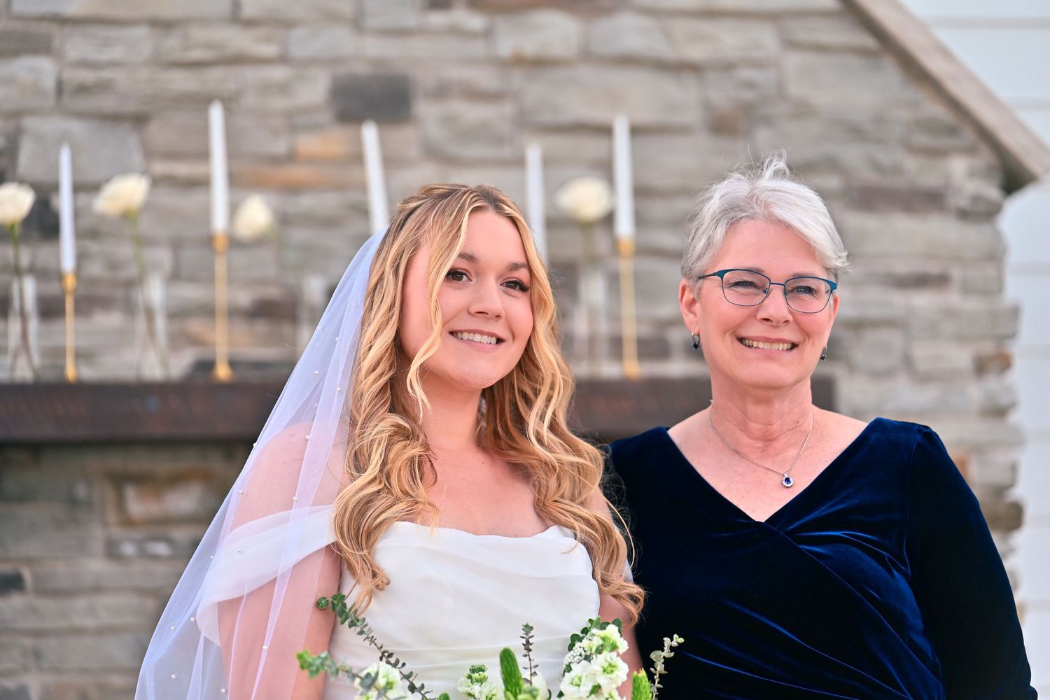 Bride in white gown and veil holds bouquet beside older woman in blue velvet dress at elegant wedding ceremony.