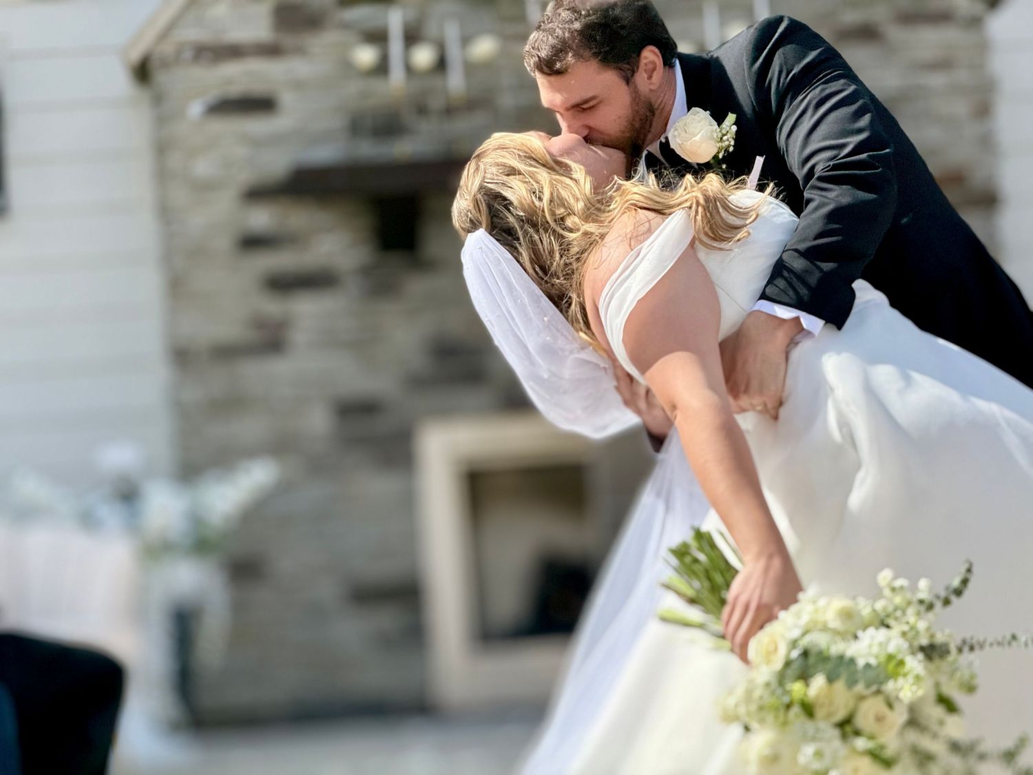 Groom dips bride for a romantic kiss in front of a stone fireplace at an elegant outdoor wedding ceremony.