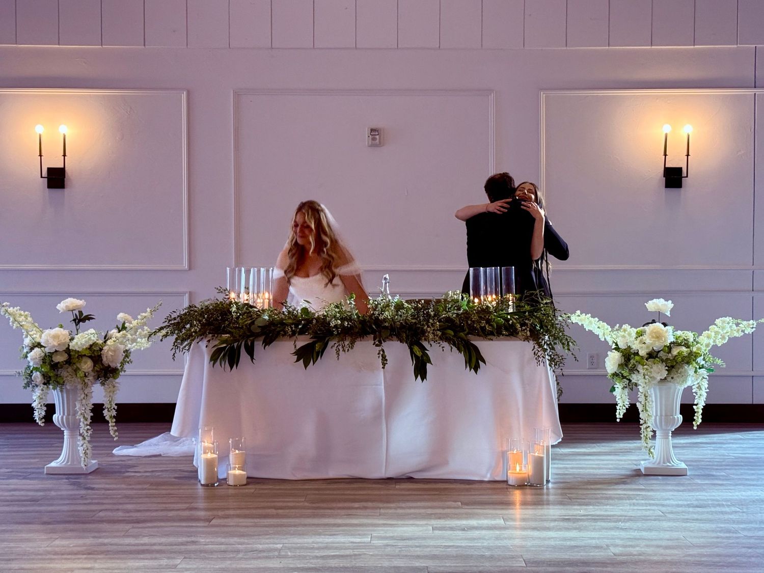 Bride and groom seated at elegant sweetheart table adorned with greenery garland and white floral arrangements in ballroom.