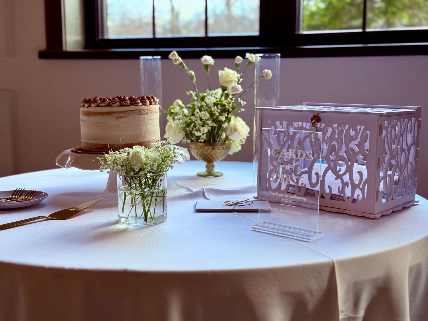 Elegant wedding reception table with white floral arrangements, tiered cake, and decorative card box by a sunlit window.