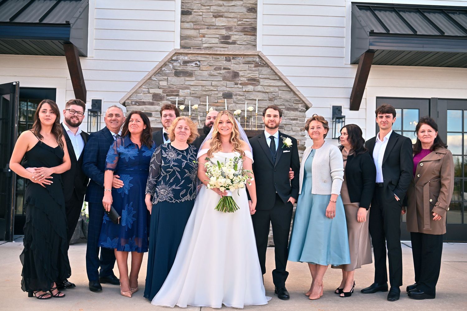 Wedding party poses outside a modern venue, bride in white gown holding bouquet, groom in suit, surrounded by family.