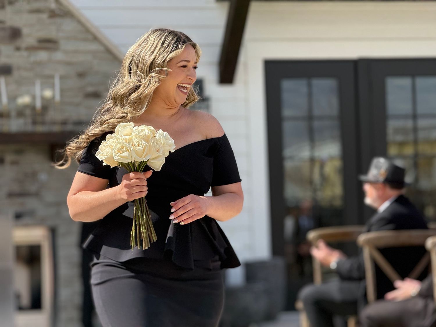 Woman in black off-shoulder dress holding white rose bouquet smiling outdoors at a wedding venue.