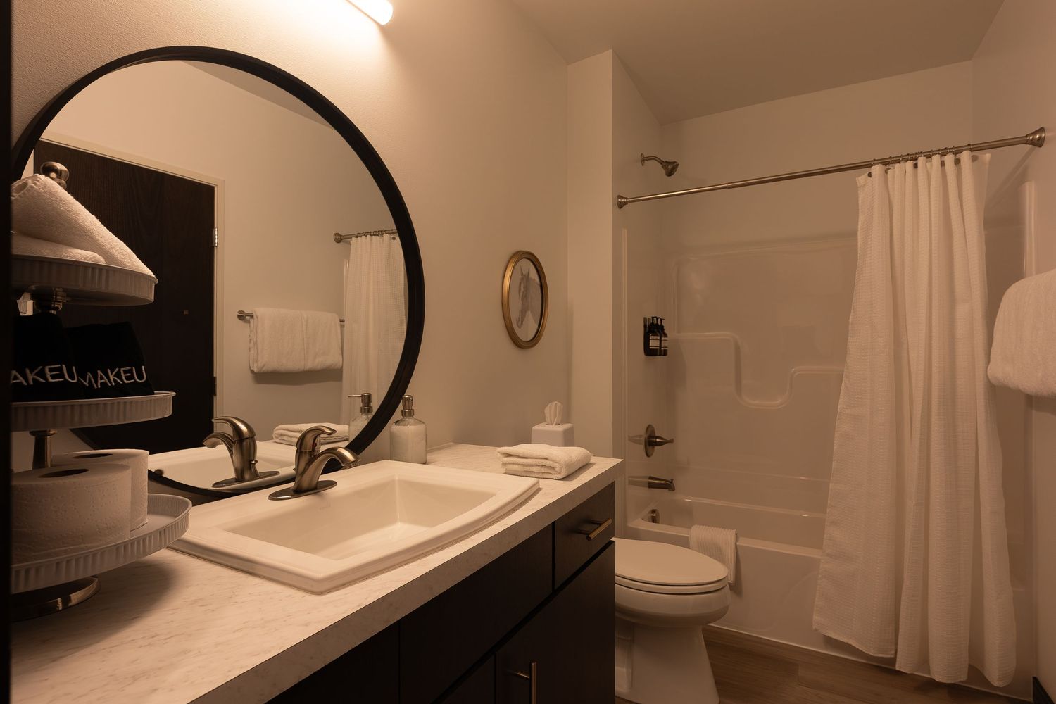 Modern bathroom interior with dark vanity, round mirror, white shower curtain and beige walls in warm lighting.