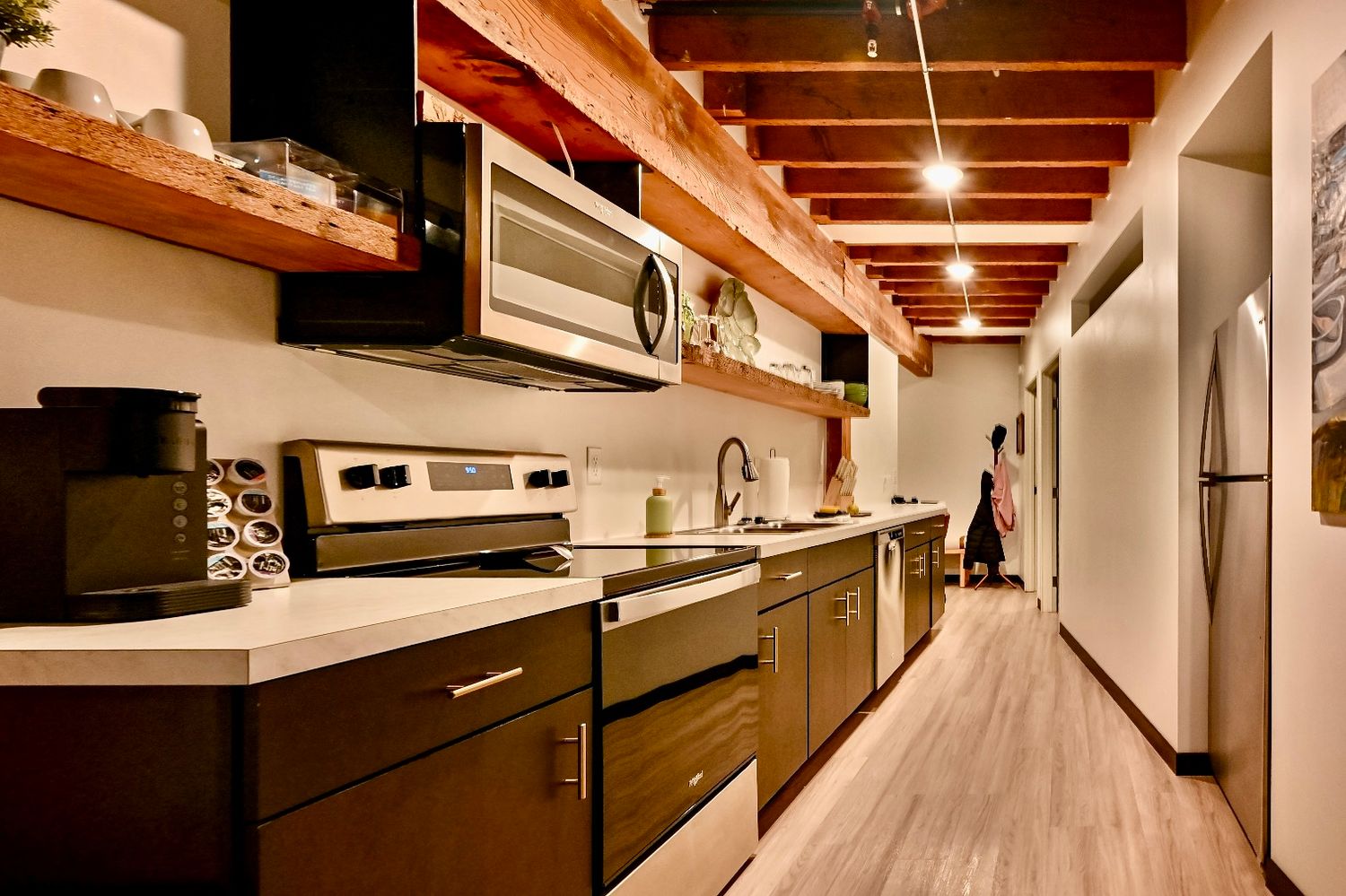 Modern galley kitchen with dark wood cabinets, white countertops, and exposed wooden beams along a narrow hallway.