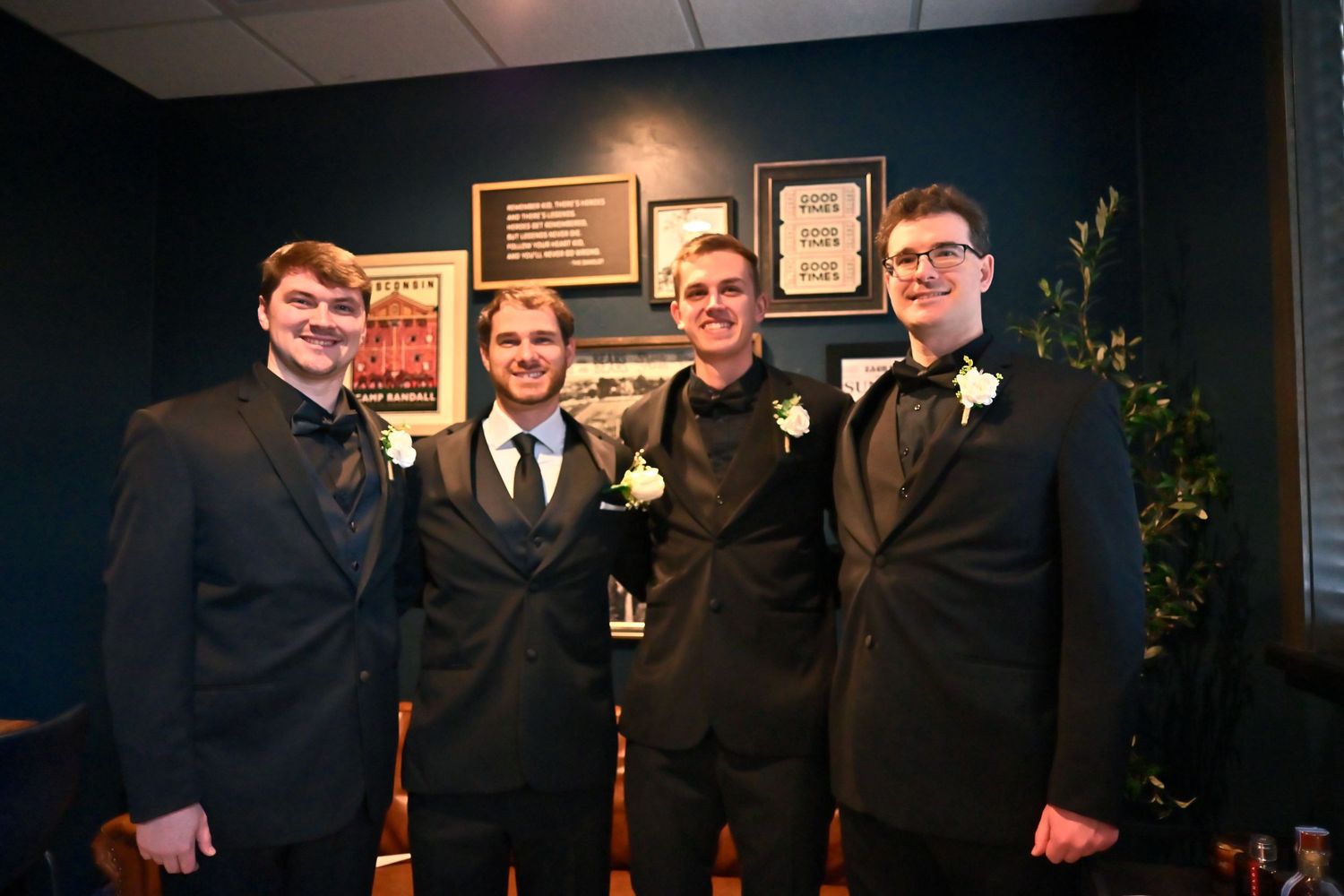 Four young men in black tuxedos with white boutonnieres smile together at a formal event.