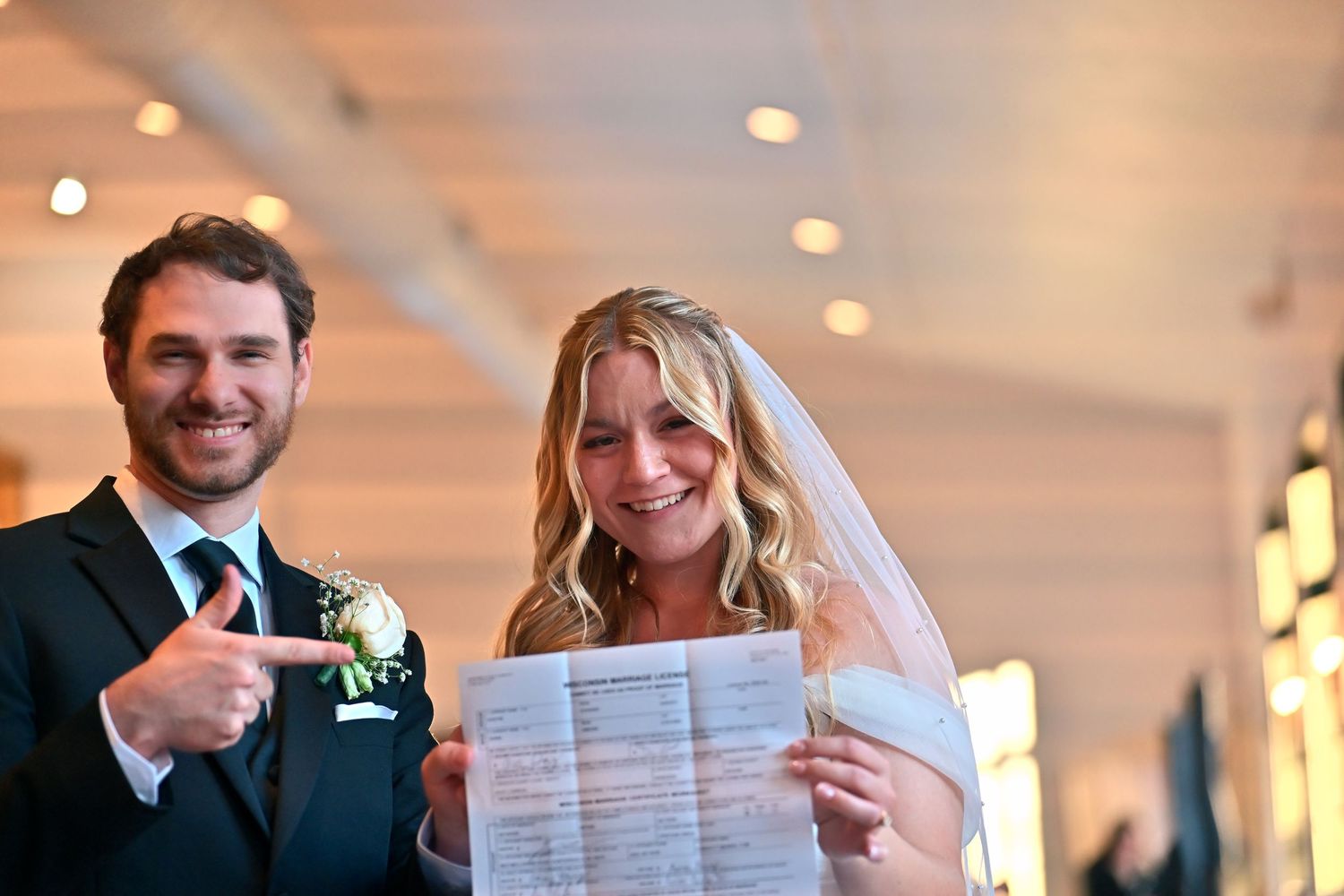 Newlywed couple smiling and holding their marriage certificate, bride in veil, groom in suit pointing at document.