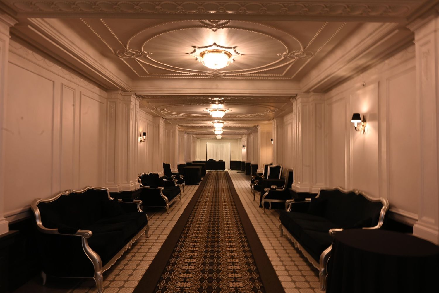 A long elegant hotel corridor with ornate ceiling, wall sconces, and dark velvet chairs lining both sides.