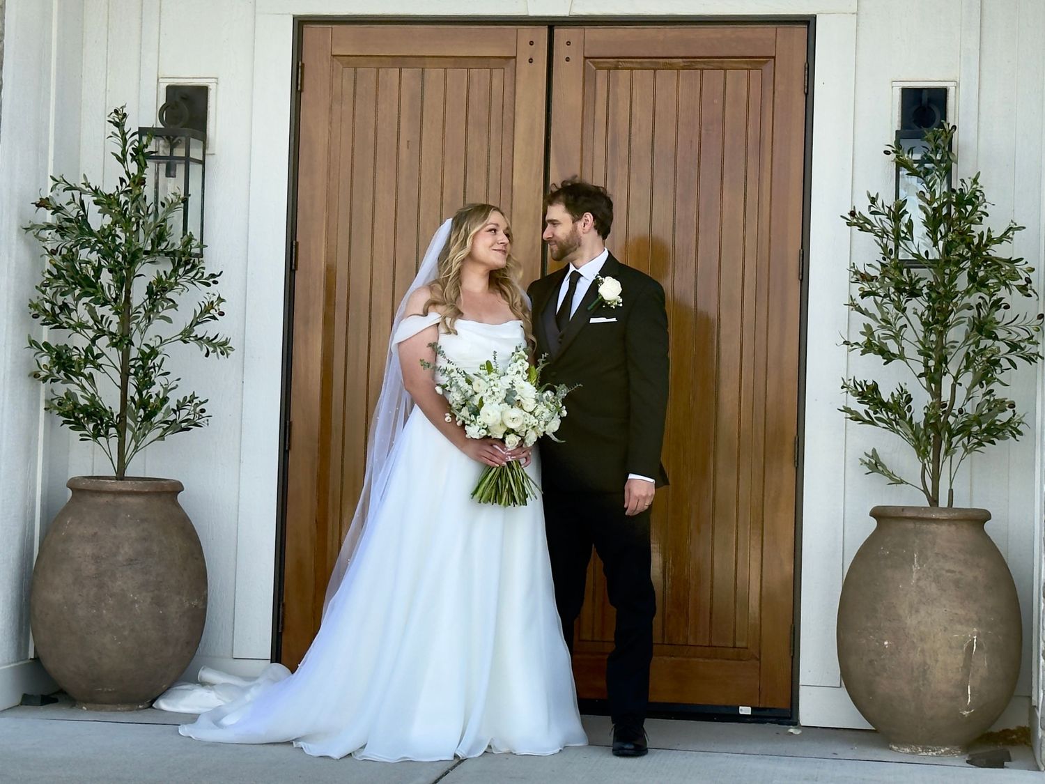 Bride and groom standing together in front of large wooden doors, flanked by olive trees in terracotta pots.