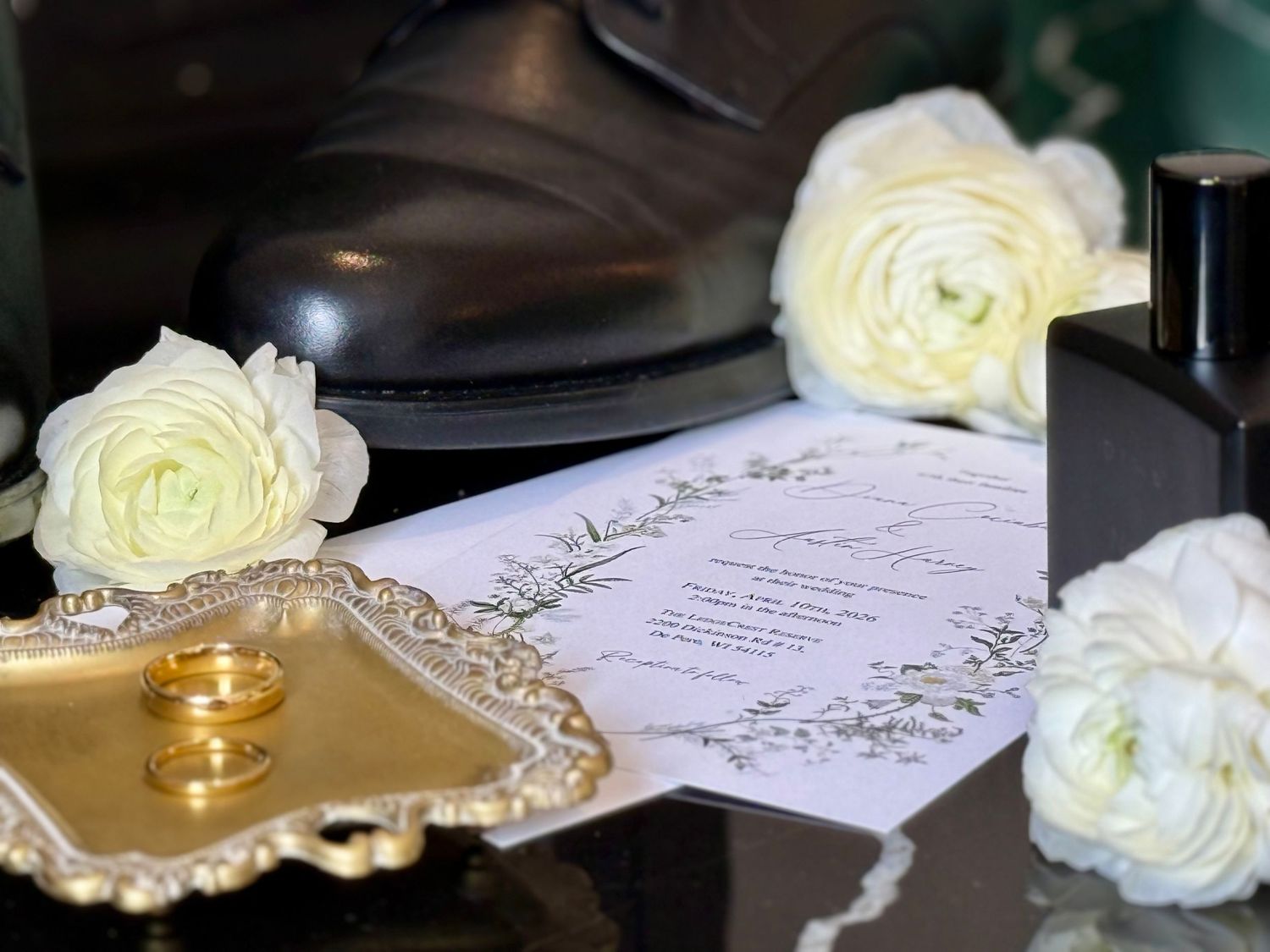 Wedding flat lay with gold rings on tray, white ranunculus flowers, dress shoes, and elegant invitation card.