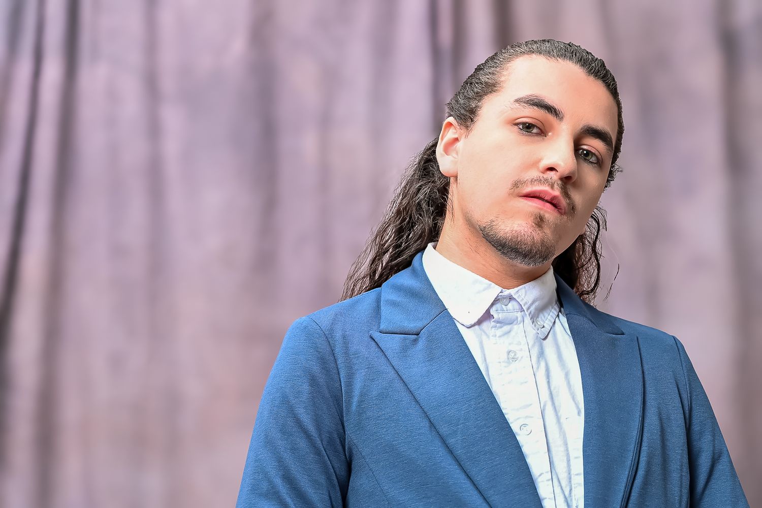 A person in a blue blazer and white shirt poses against a purple curtain backdrop with long hair styled back.
