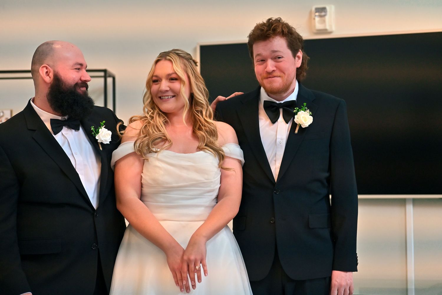 Bride in white gown smiles with two men in tuxedos at elegant wedding reception venue.