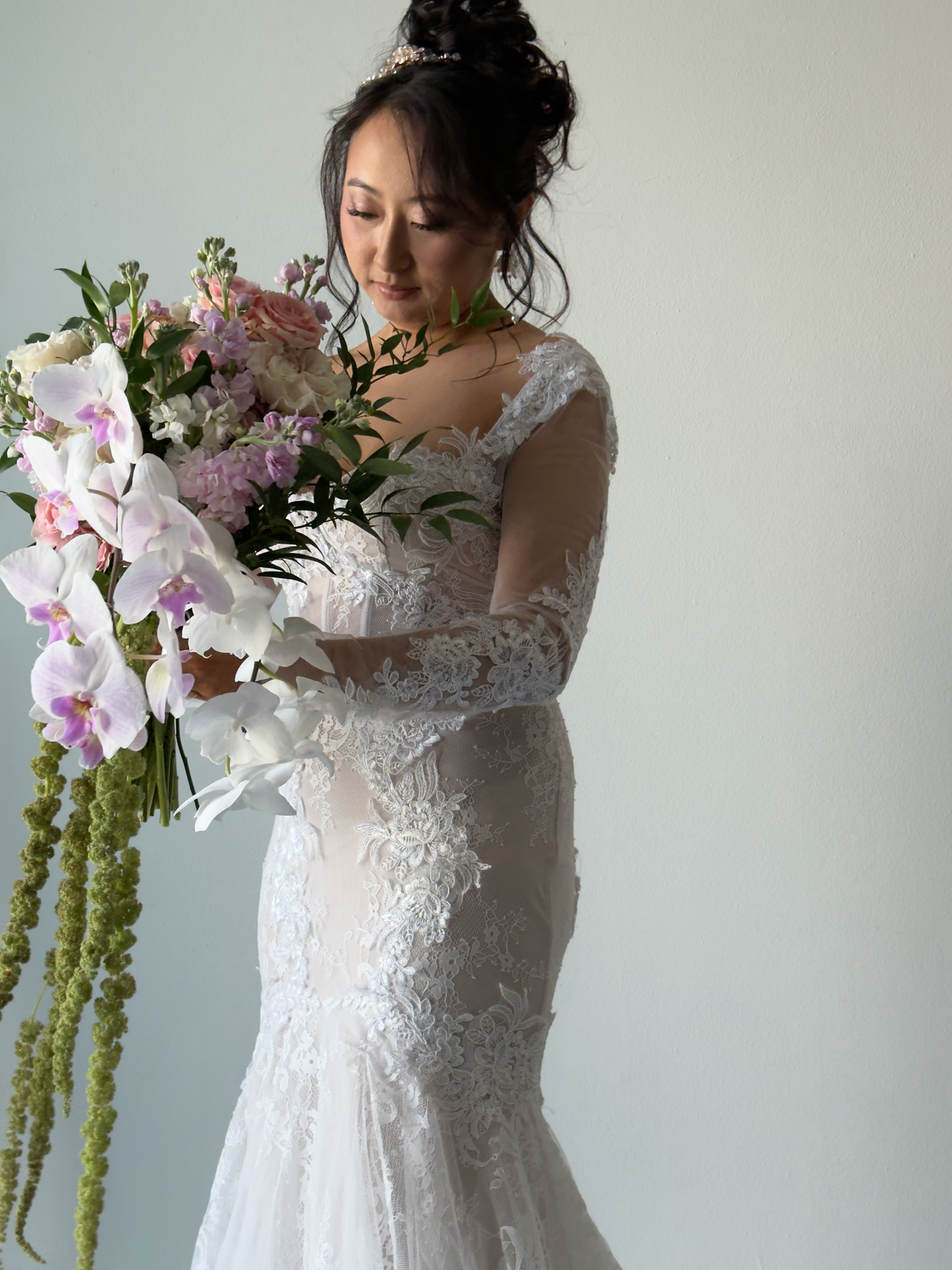 A bride in a delicate lace wedding dress poses with elegant pink and white orchids and green hanging amaranthus.