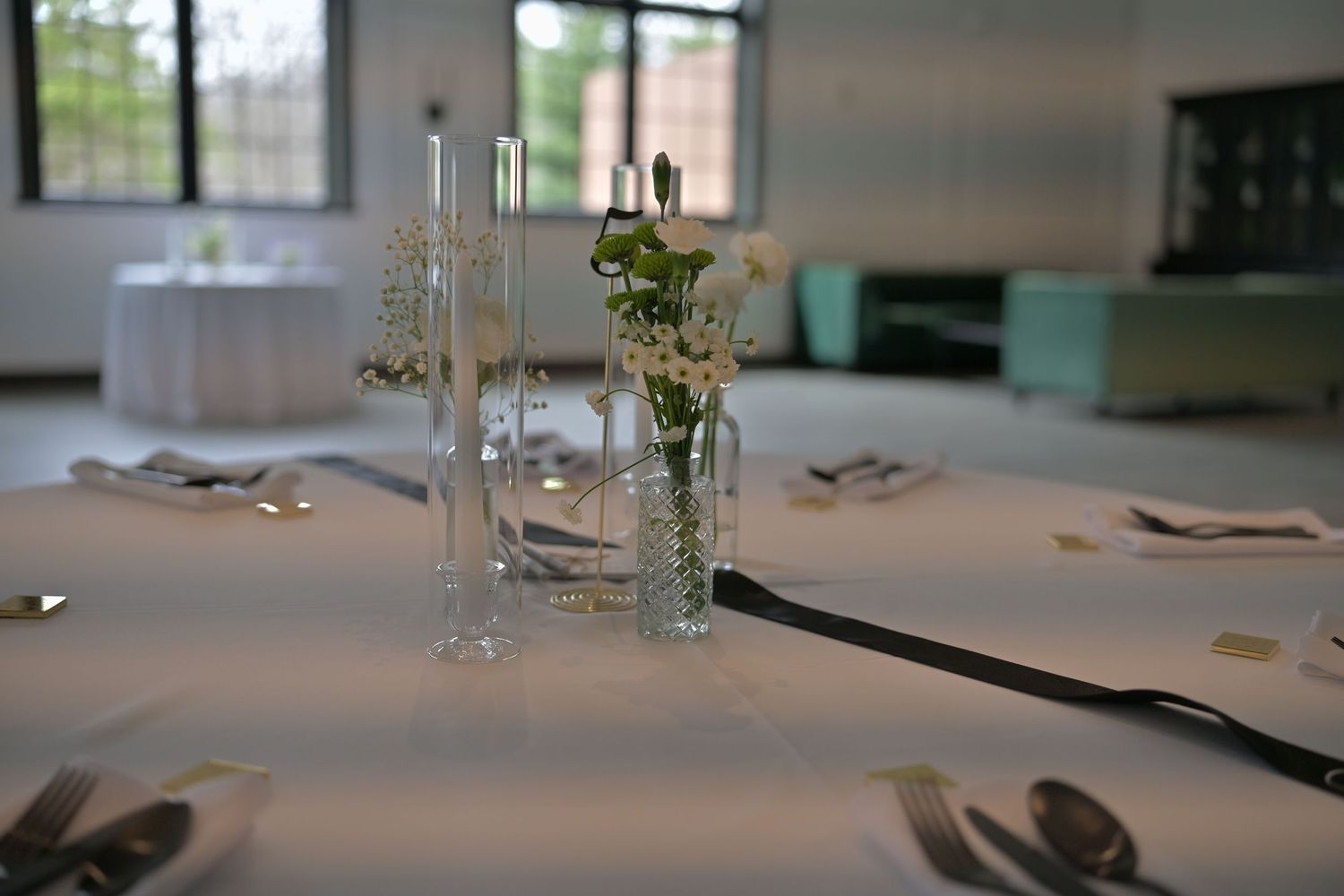 Elegant wedding reception table setting with glass vase centerpiece featuring white flowers and greenery in a bright venue.