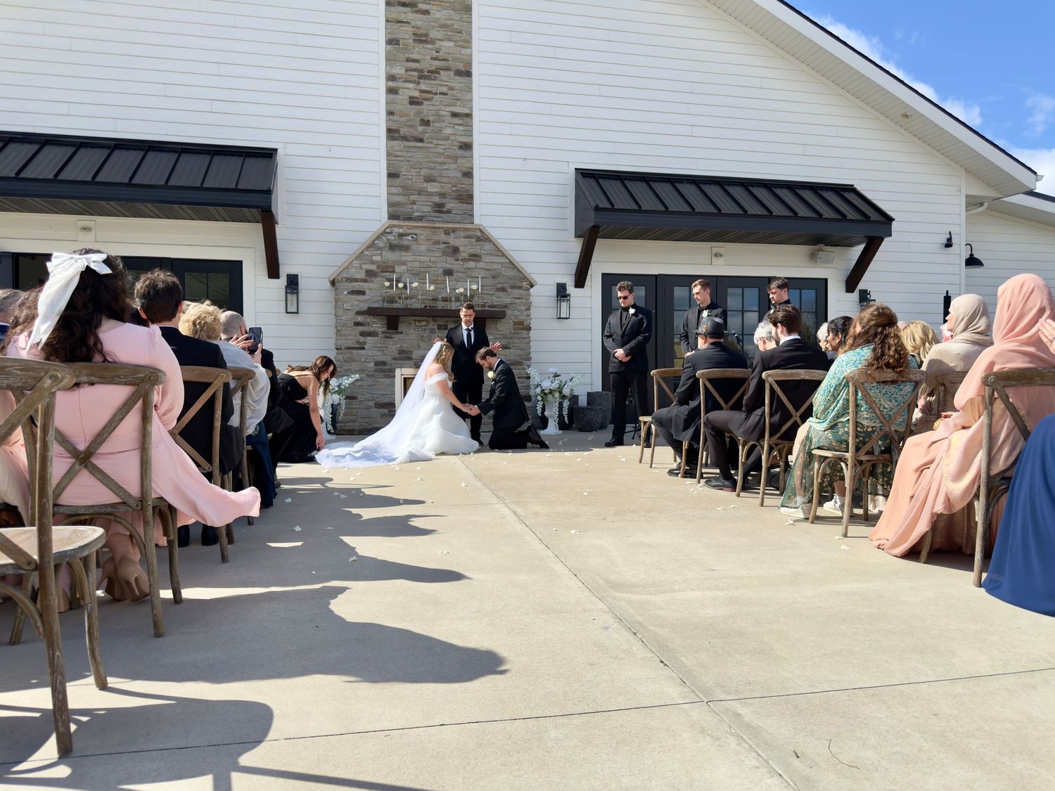 Outdoor wedding ceremony at a white barn venue with guests seated on both sides as couple exchanges vows.