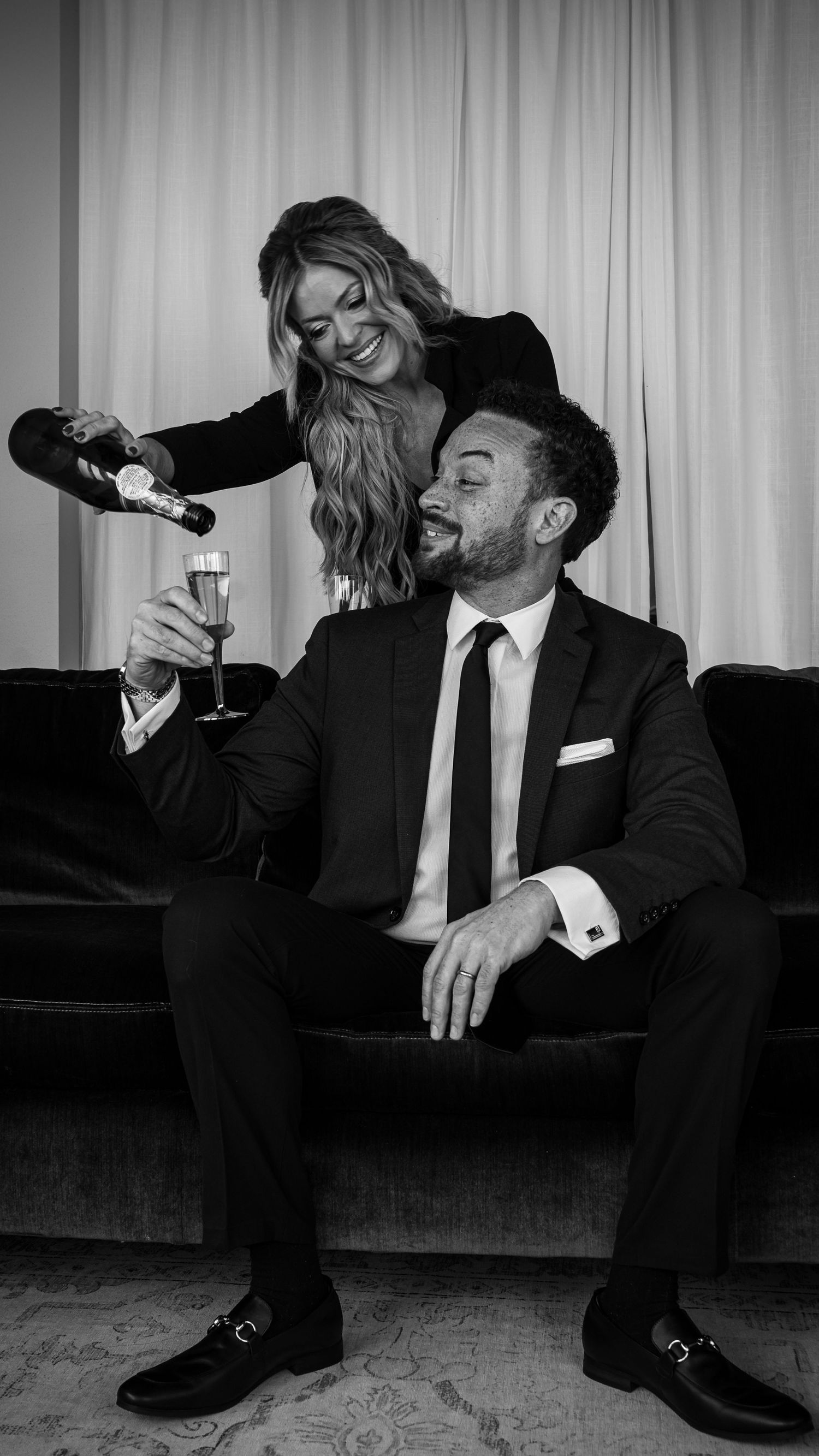 Black and white photo series showing a couple in formal attire laughing and sharing champagne on a couch.