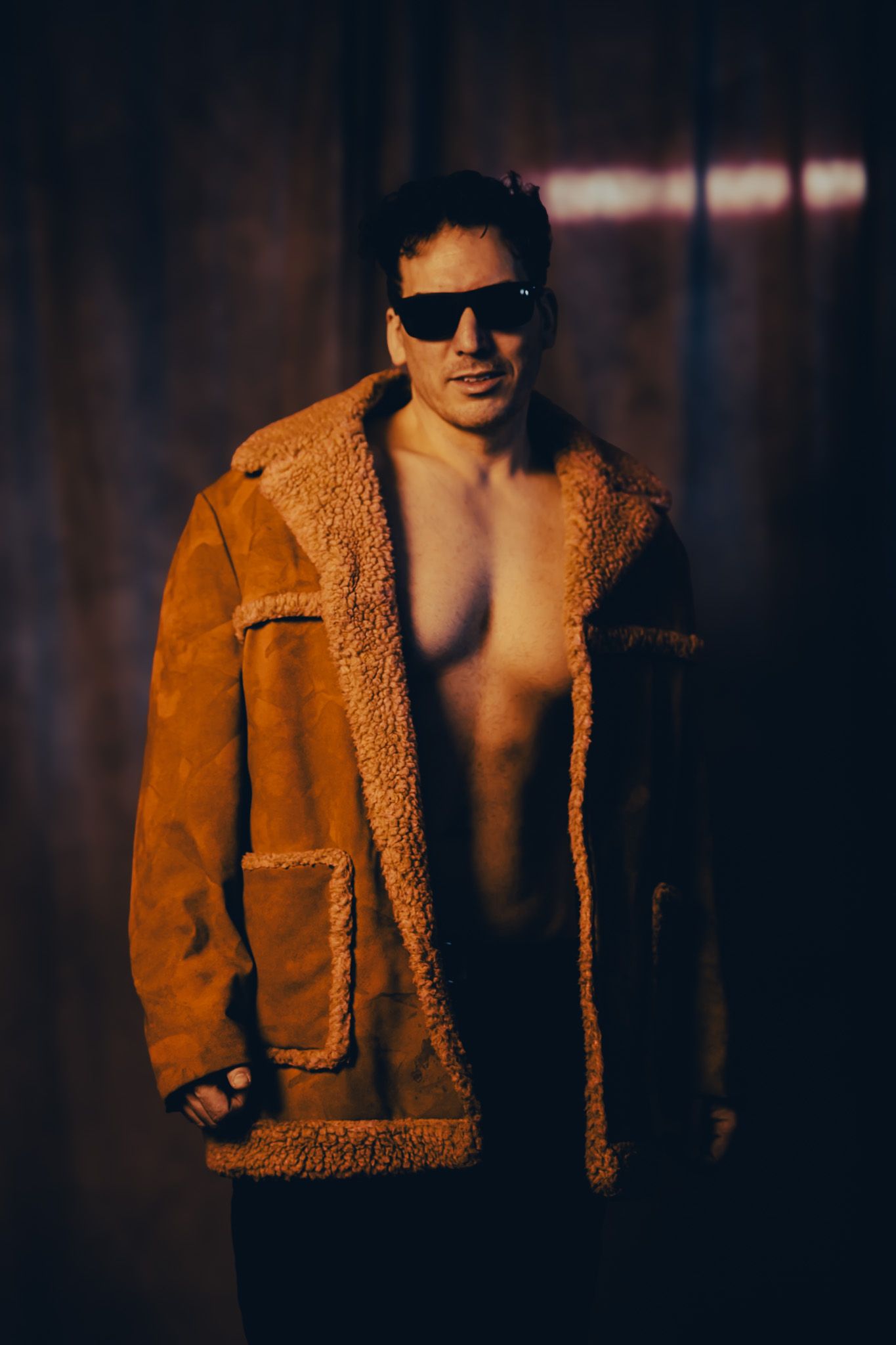 Shirtless person wearing sunglasses and brown shearling coat poses against dark background in moody lighting.