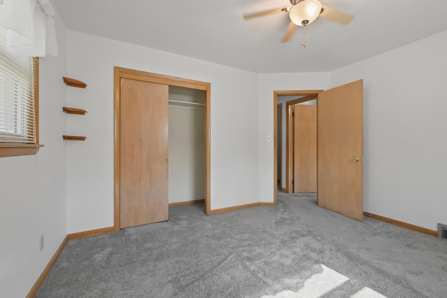 Spacious empty bedroom with beige carpet, ceiling fan, and white walls showing multiple doorways and natural lighting.