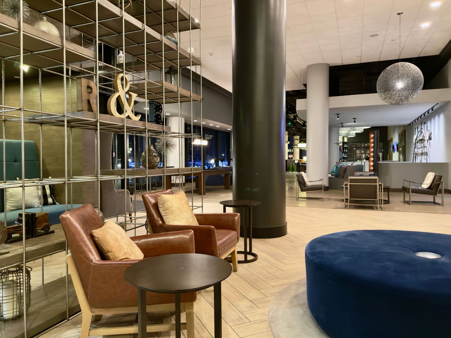 Modern hotel lobby interior with brown leather chairs, blue ottoman, and metal shelving display against dark columns.