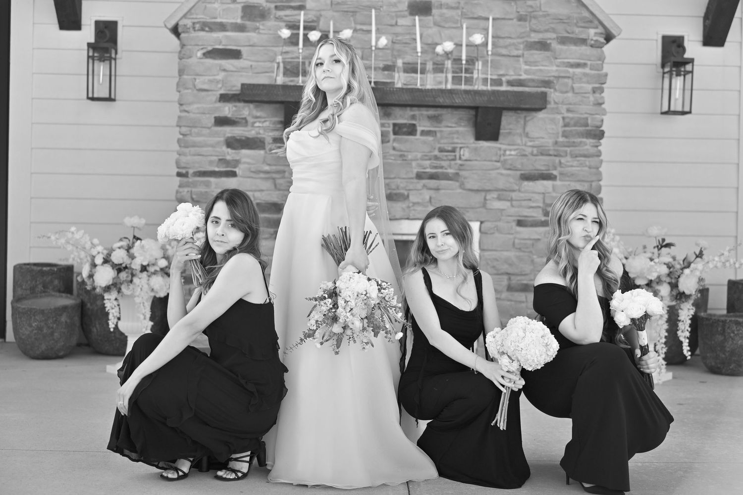 Black and white wedding photo of bride in white gown with three bridesmaids in black dresses posing near stone fireplace.