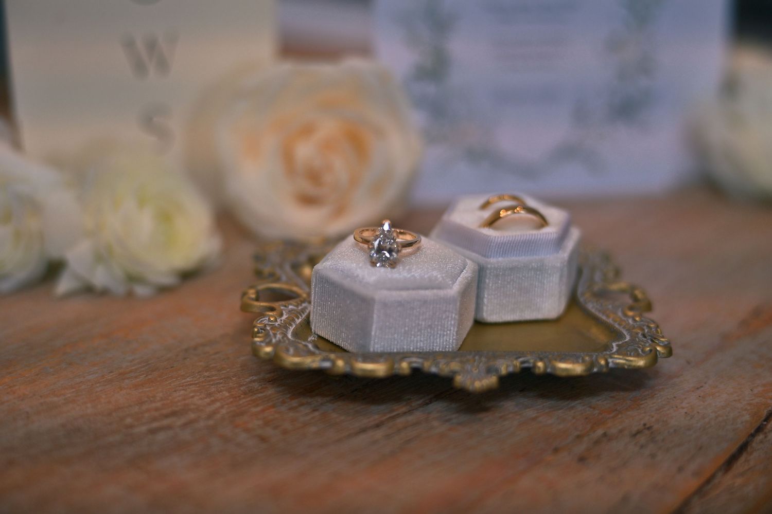 Two wedding rings displayed in velvet hexagon boxes on a gold tray, surrounded by white roses and elegant stationery.
