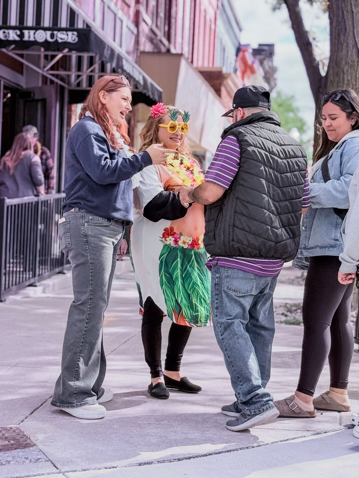 A group of people gather on a sidewalk, with one wearing a festive Hawaiian-style grass skirt.