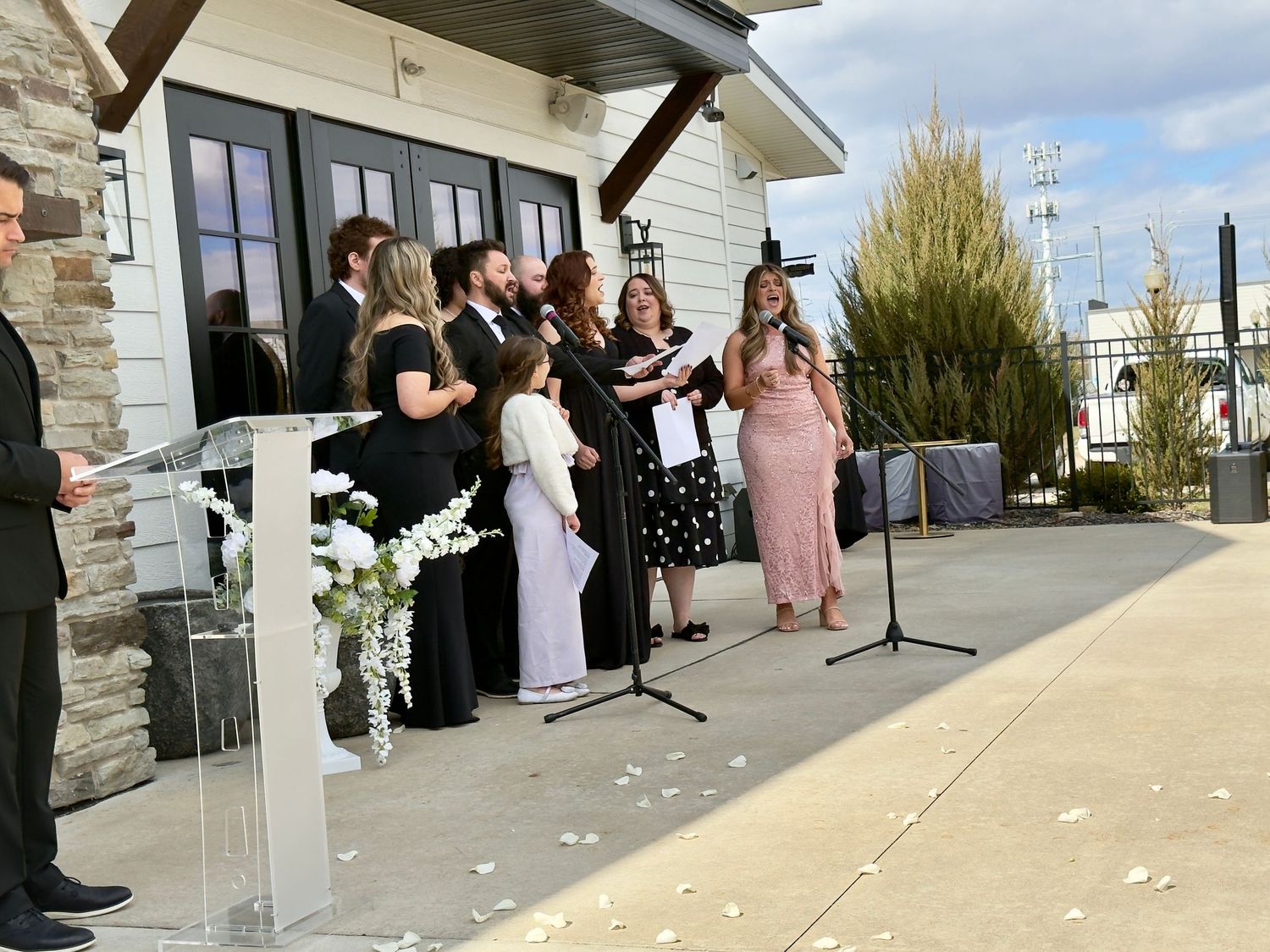 Wedding ceremony outdoors with bridal party and guests gathered near a podium adorned with white flowers.