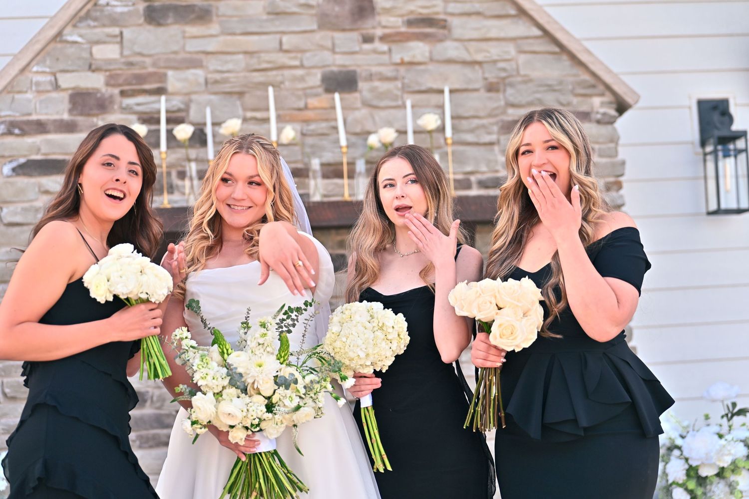 Bride in white gown with three bridesmaids in black dresses holding white bouquets, laughing outdoors.