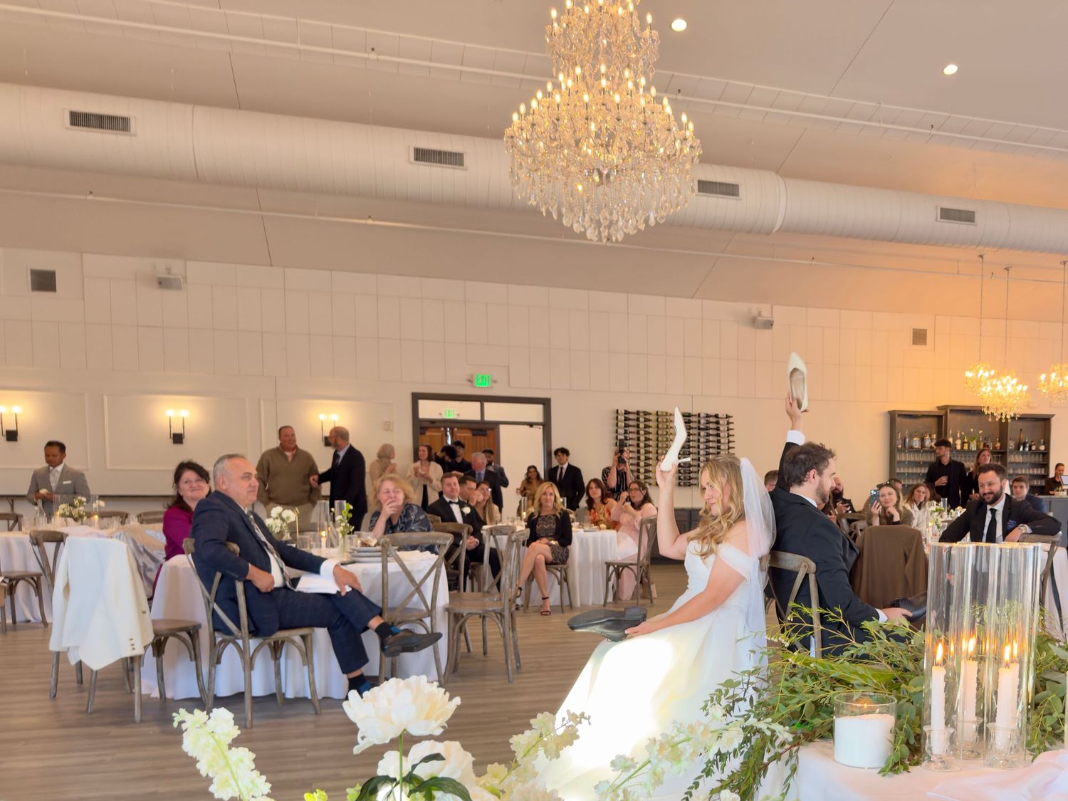 Elegant wedding reception in a bright venue with crystal chandelier, guests seated at tables, bride and groom at head table.
