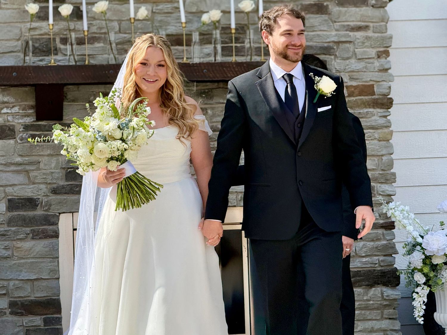 Bride in white gown holding bouquet walks with groom in navy suit outside stone church on wedding day.