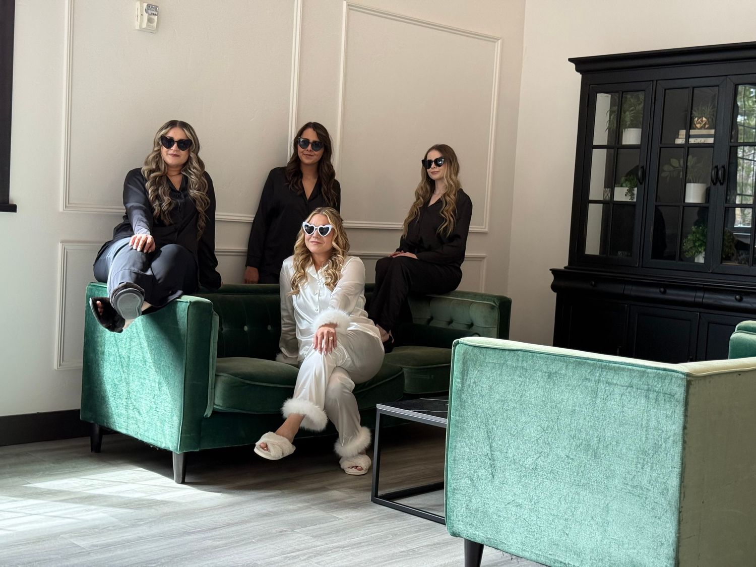 Four stylish women wearing sunglasses pose on green velvet chairs in a modern, elegant lounge setting.