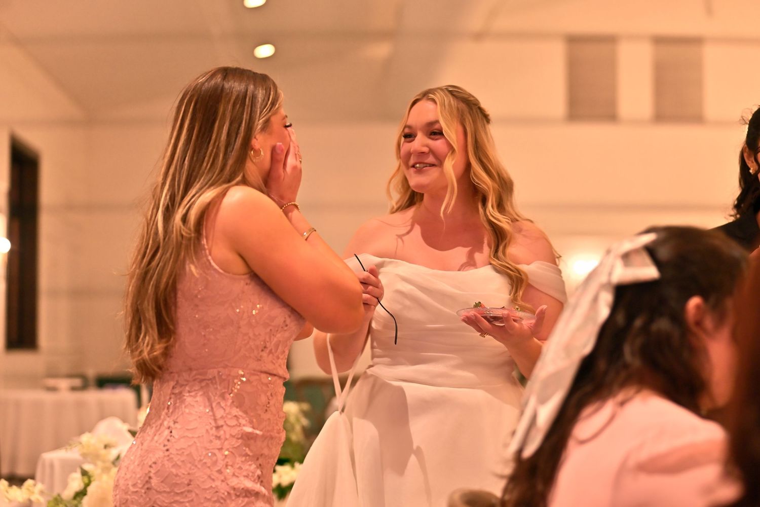 Bride in white dress sharing emotional moment with guest in pink lace gown at elegant wedding reception.