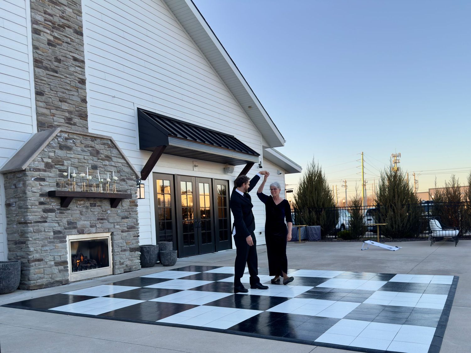 Couple dancing on black and white checkered outdoor dance floor at elegant venue with stone fireplace at sunset.
