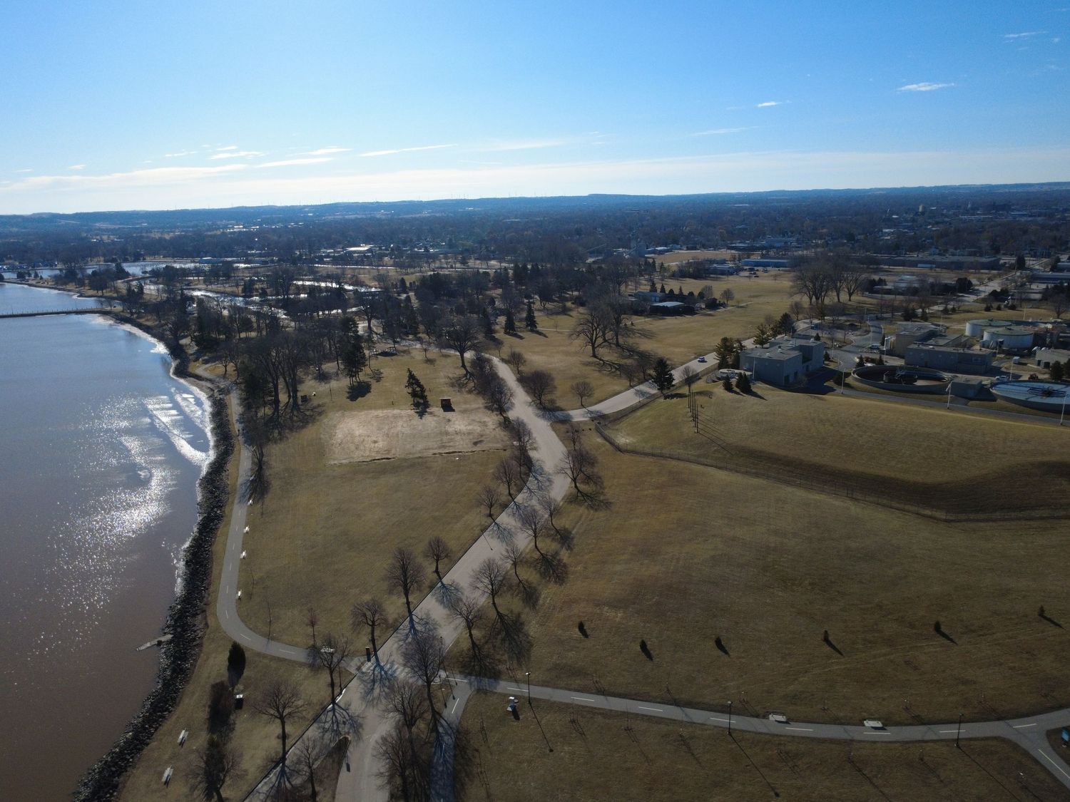 Aerial view of a coastal park with tree-lined paths and open fields next to a waterfront on a sunny winter day.