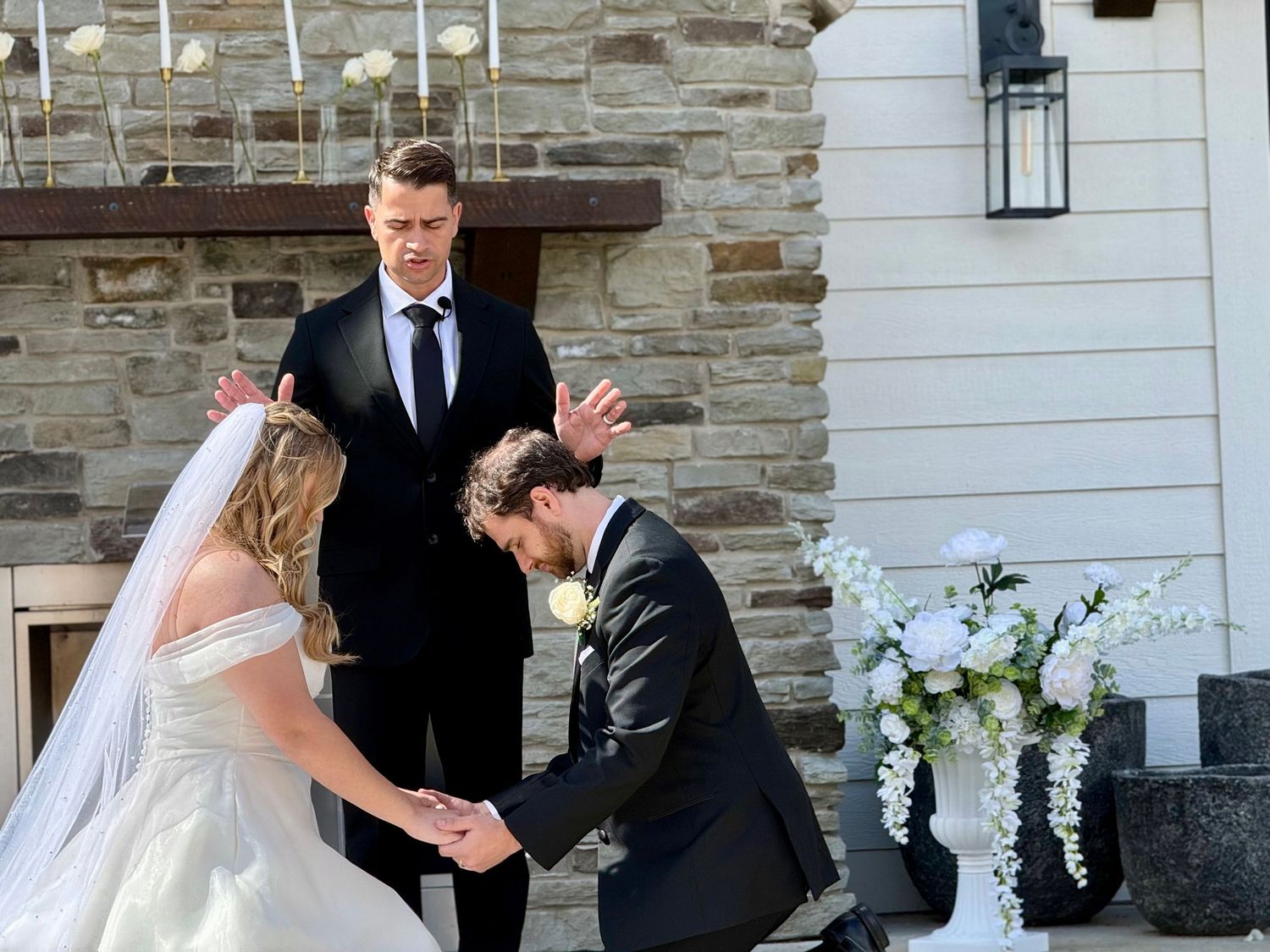 Groom kneeling before bride during outdoor wedding ceremony while officiant stands behind them near stone wall.
