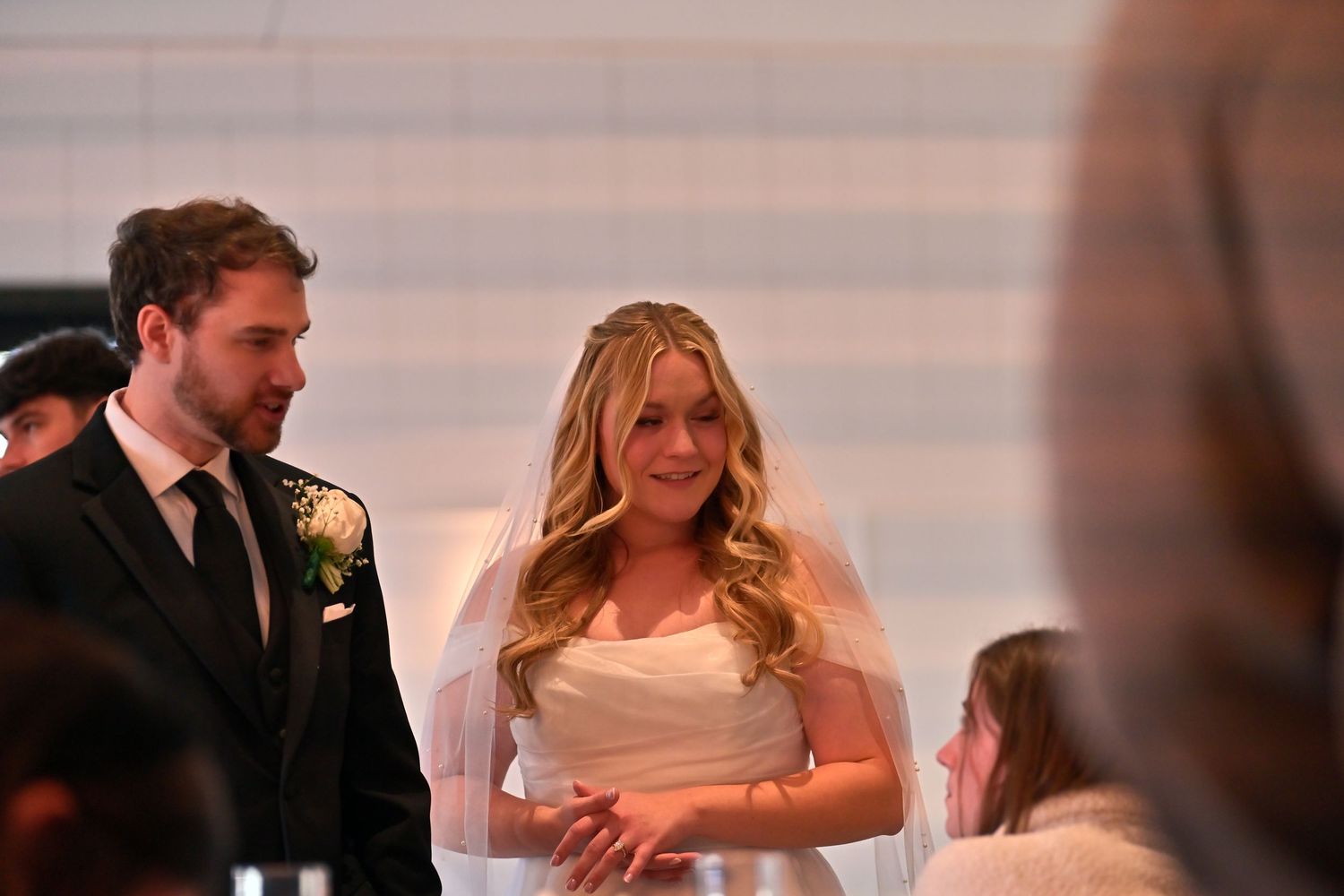 Bride in white gown and veil stands with groom in dark suit during wedding ceremony indoors.