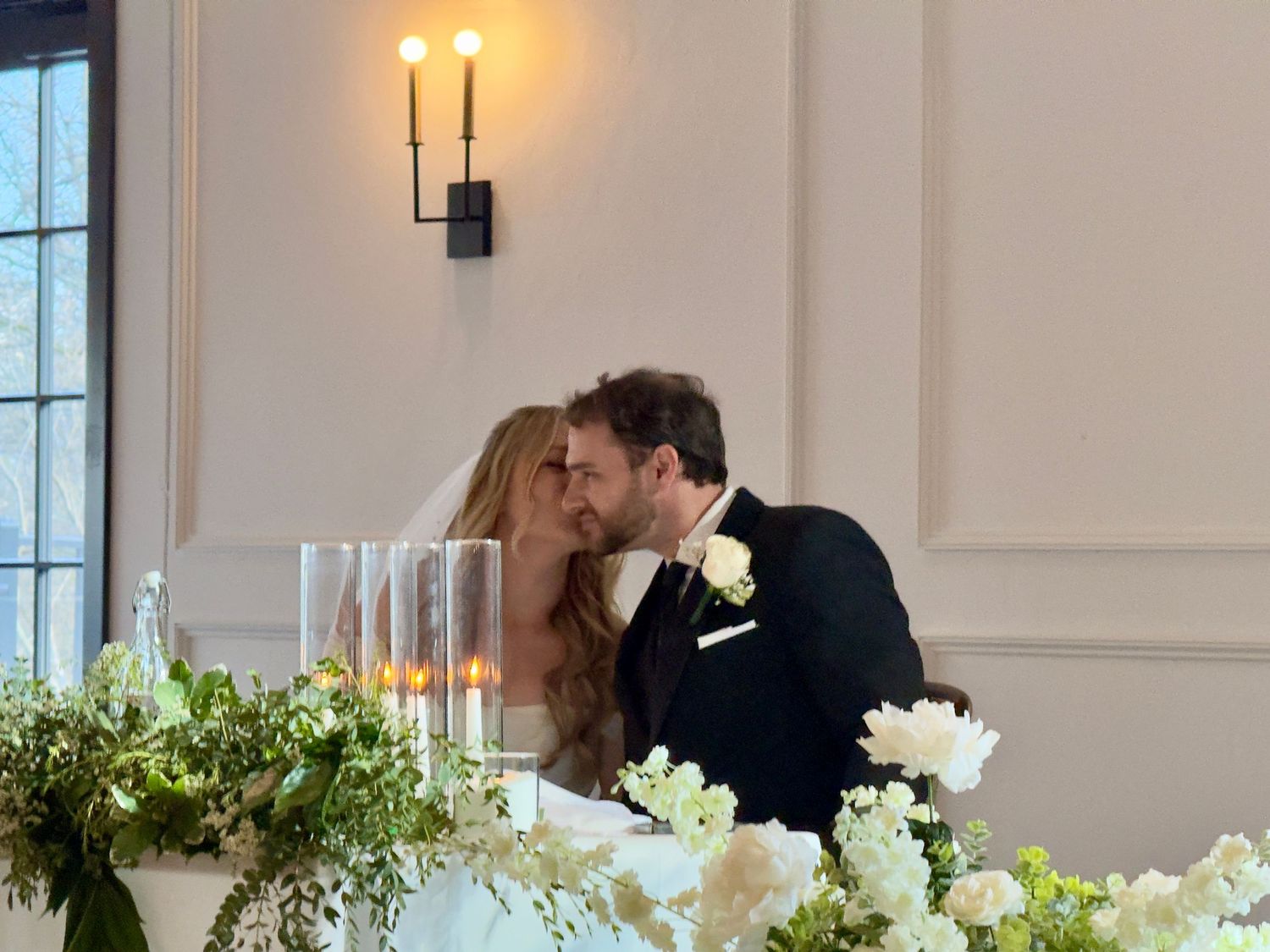 Bride and groom sharing a kiss at wedding reception sweetheart table adorned with white flowers and candles.