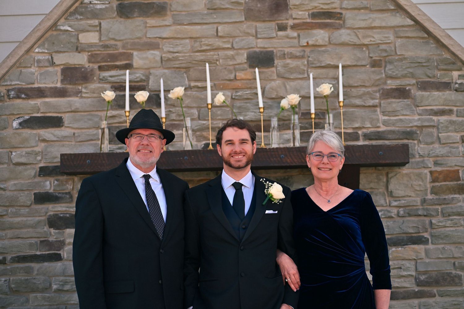 Family of three posing formally in front of stone fireplace with candles at a wedding celebration.