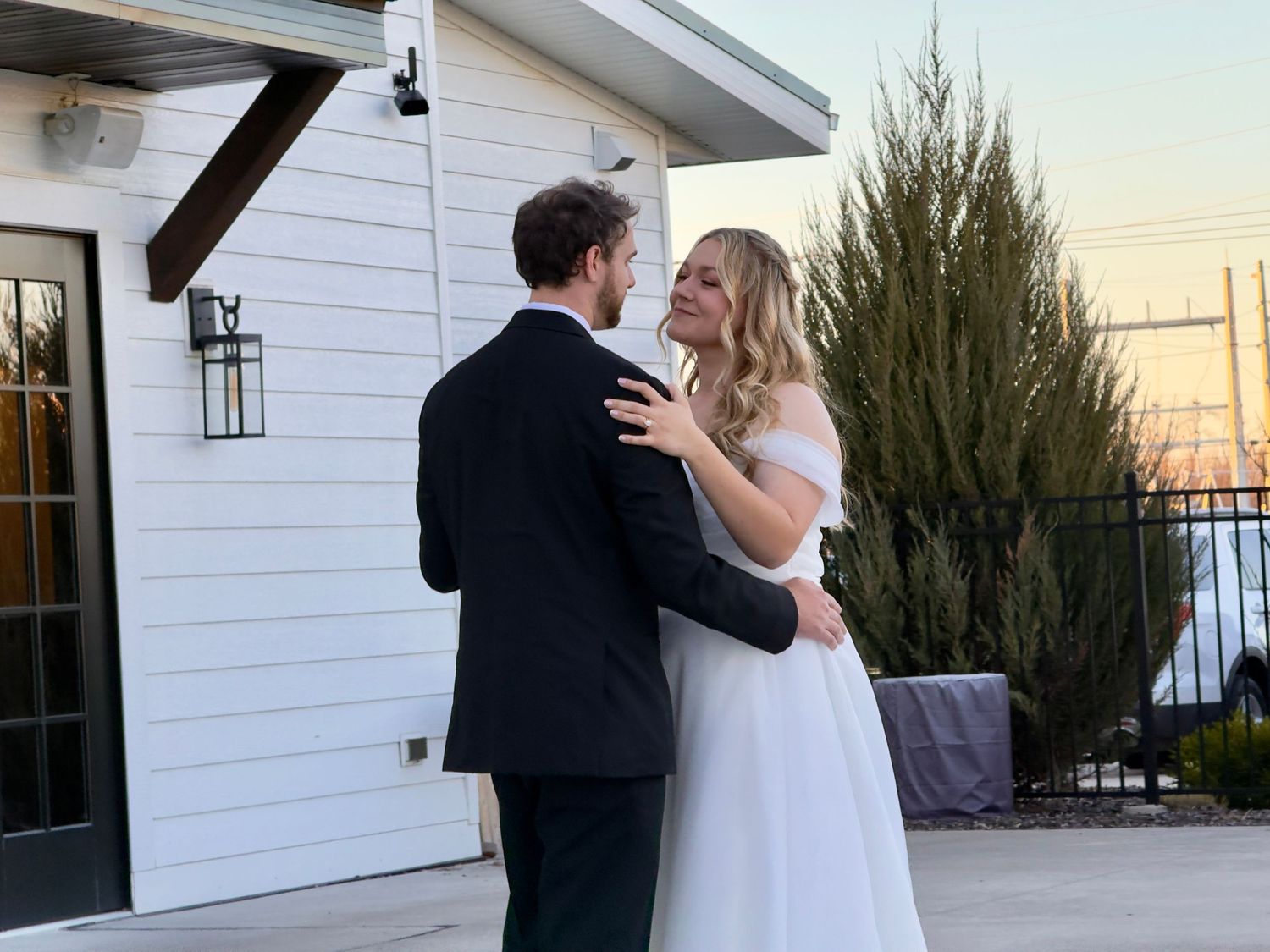 Bride and groom share a romantic first dance outdoors at dusk near a white building.