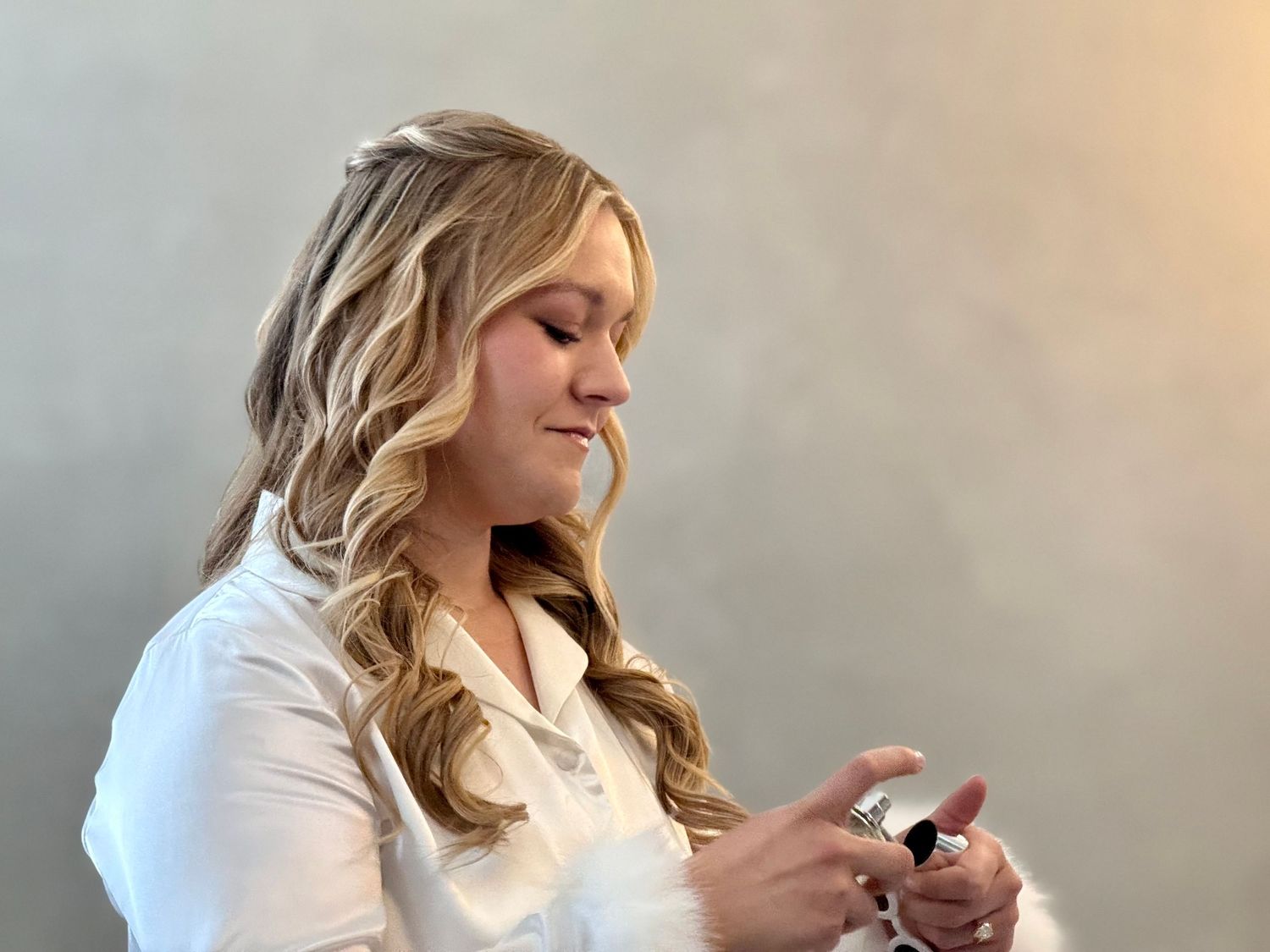 Young blonde woman in white shirt smiling while looking at her phone against a gray wall.