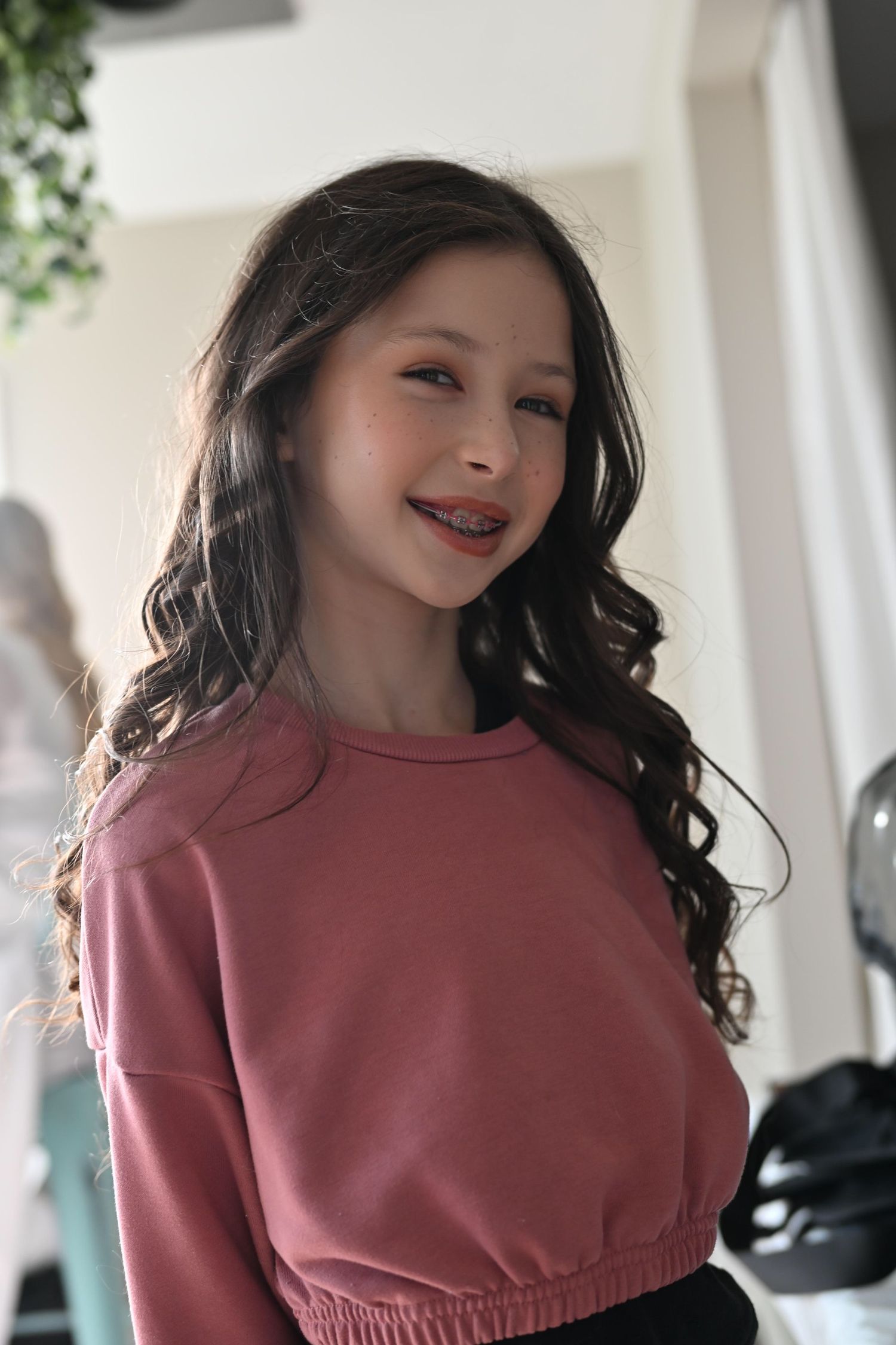 A person wearing a pink cropped sweatshirt smiles warmly while posing indoors with long dark wavy hair.