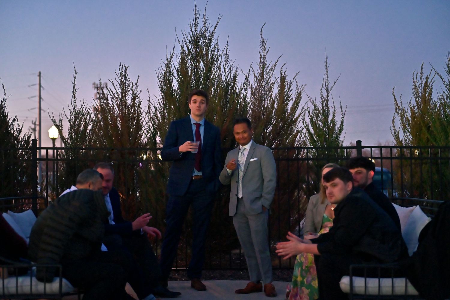 Men in suits socializing on a rooftop terrace at dusk with tall hedges and city views.