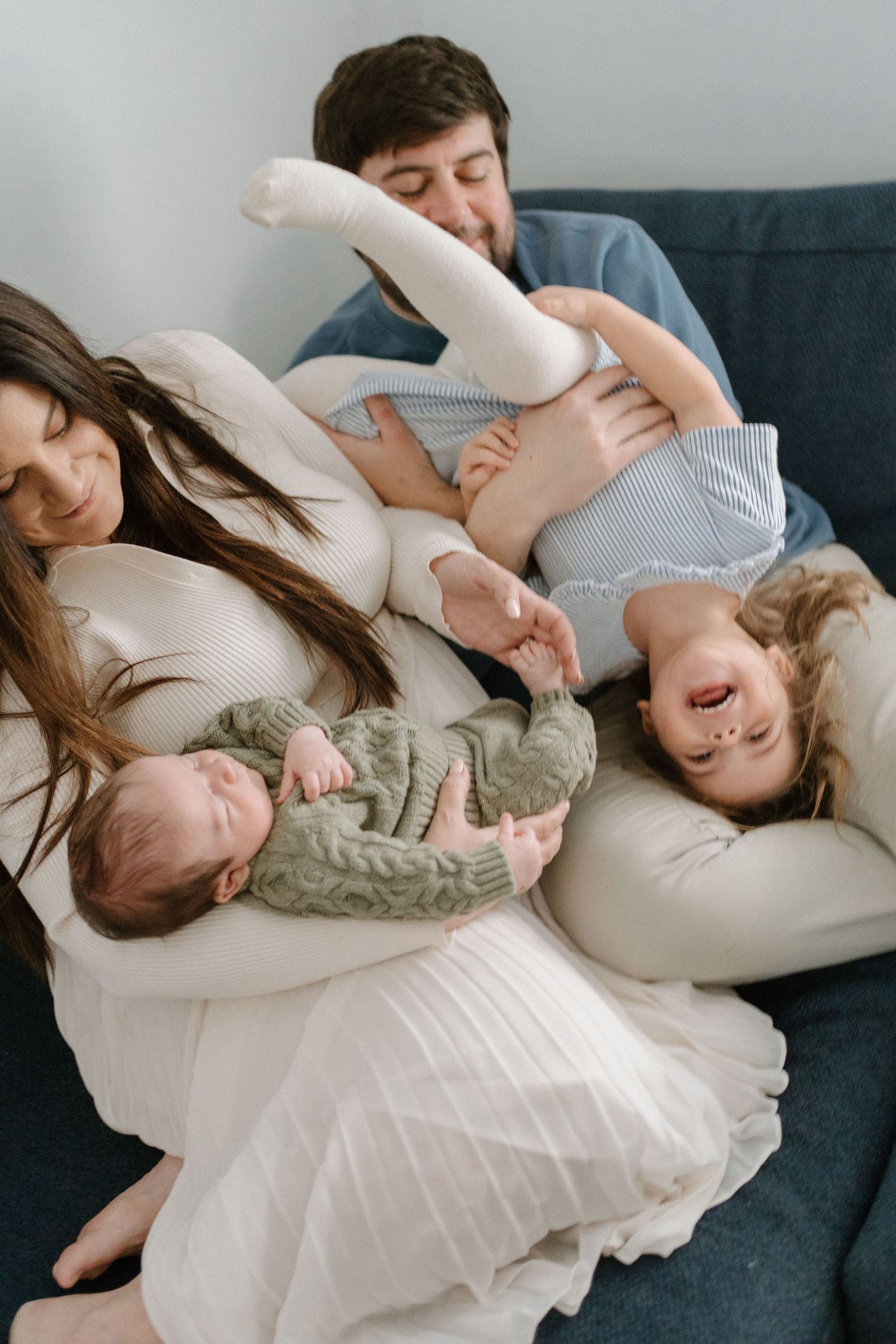 A joyful family of four plays together on a blue sofa, with parents holding their two young children.