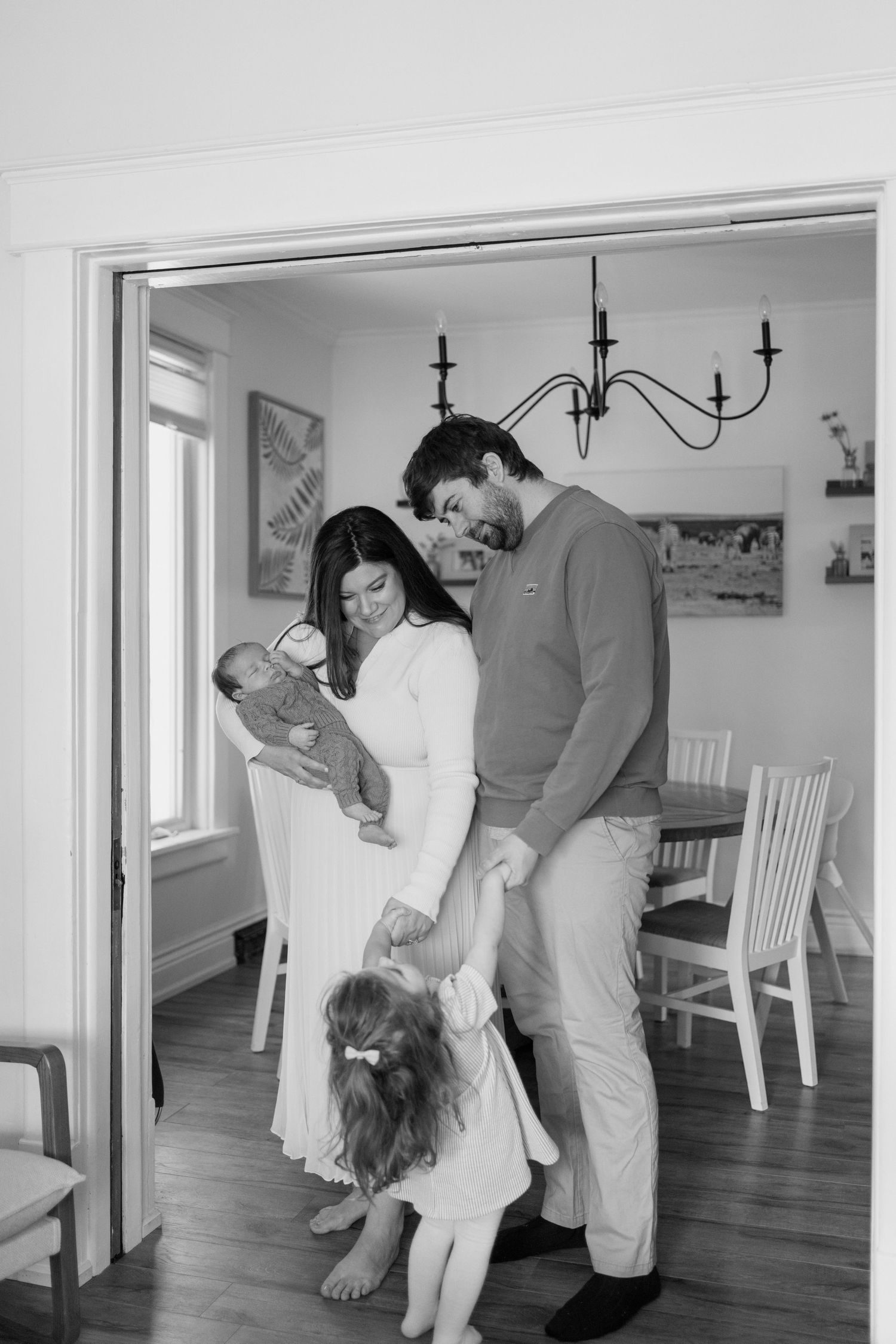 A black and white photo of a family of four standing in a doorway, parents holding newborn and toddler.