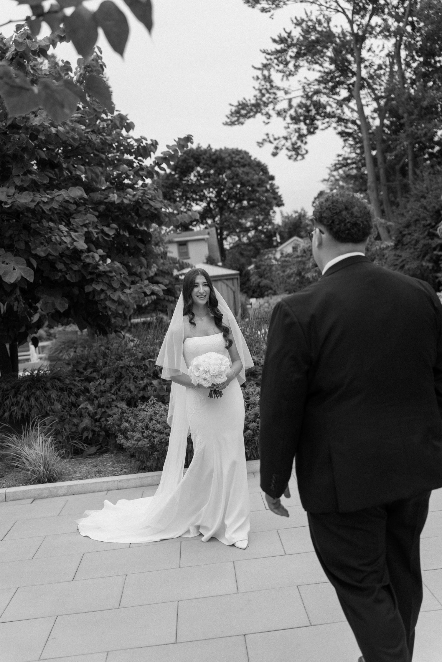 Black and white photo of a bride in a white gown holding bouquet smiling at groom in suit outdoors.