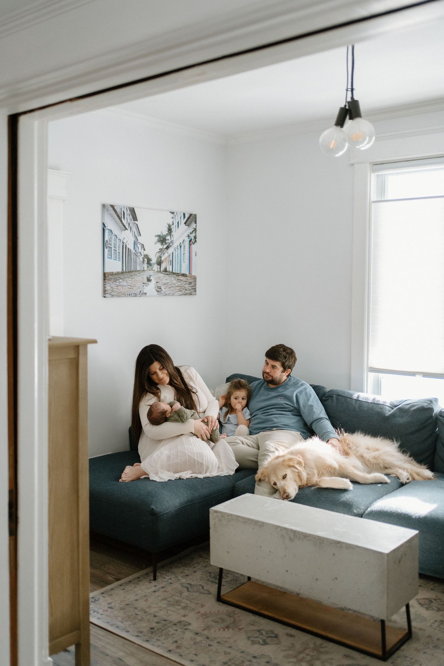 A family relaxes on a teal sofa with their newborn, toddler, and large golden dog in a bright living room.